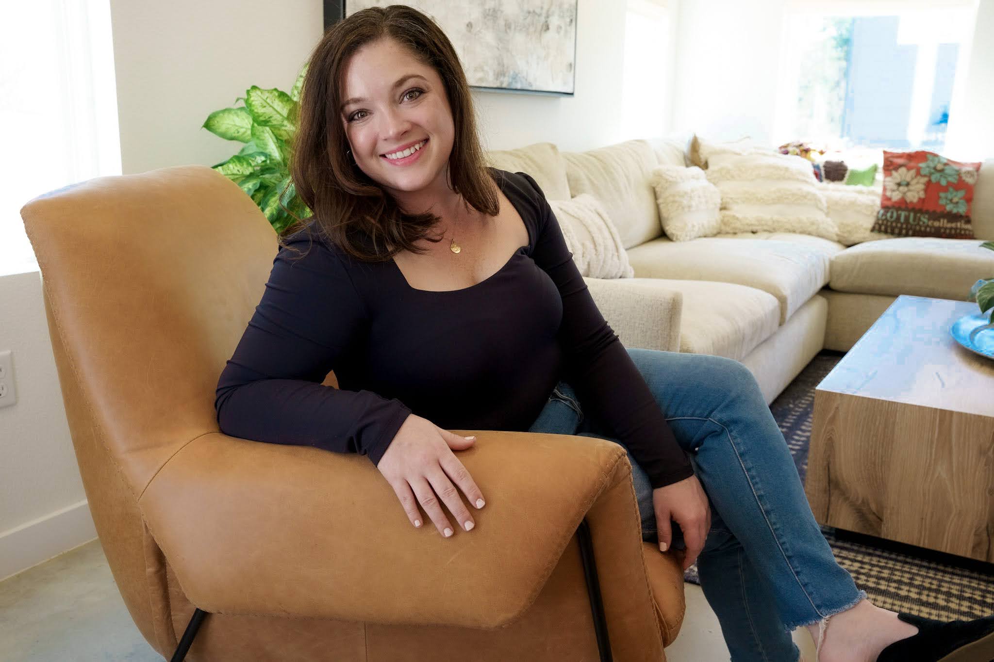Woman sitting on a tan armchair in a living room, smiling, with a sofa and window in the background.