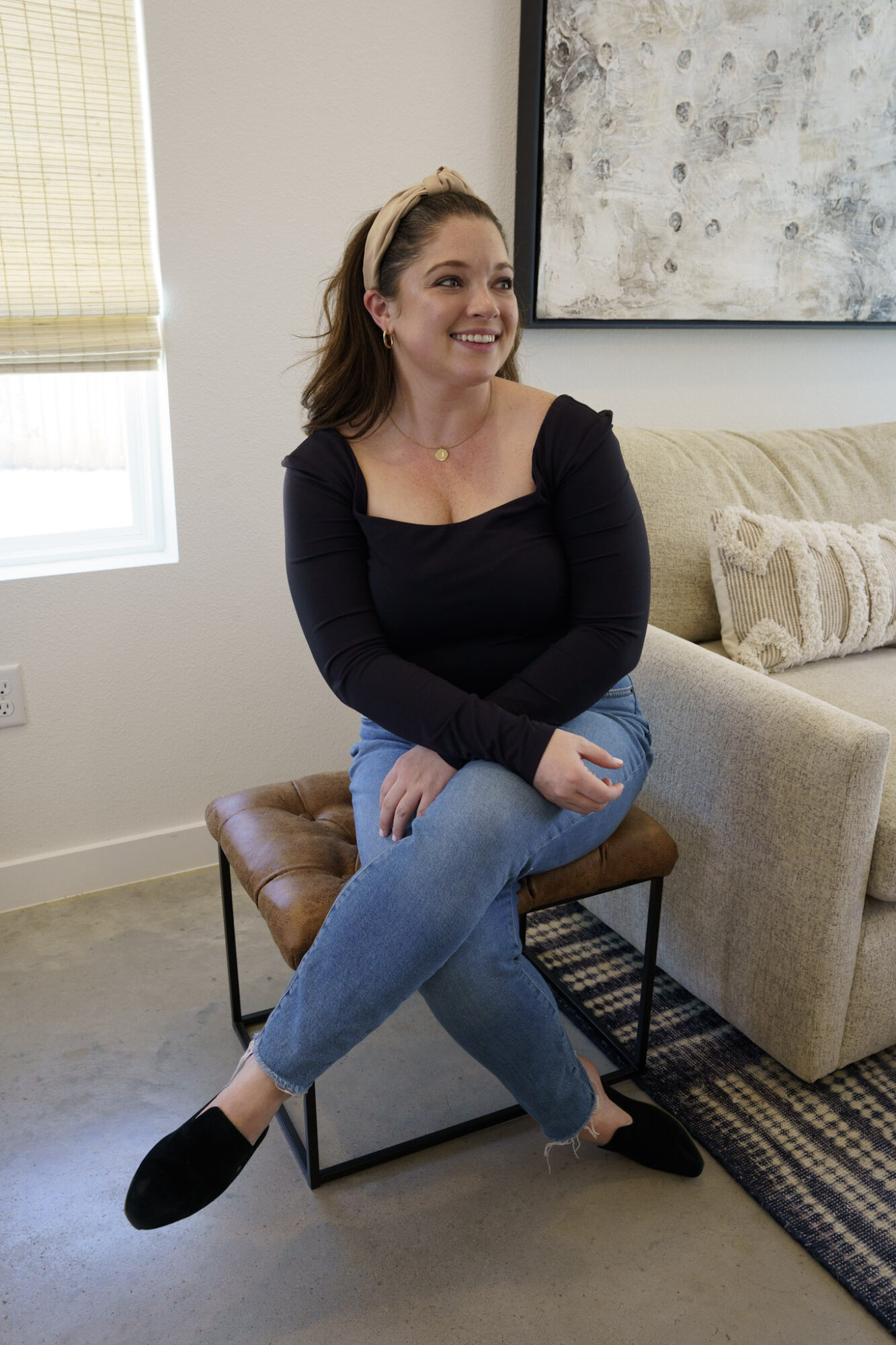Smiling woman sitting on a small brown stool in a living room, wearing a black top and blue jeans.