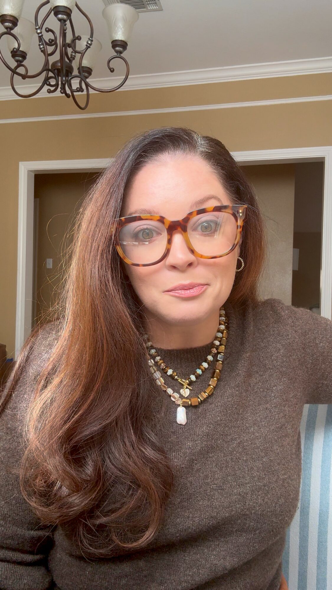 Woman with long brown hair, glasses, and layered necklaces, wearing a brown top, standing indoors near a doorway.