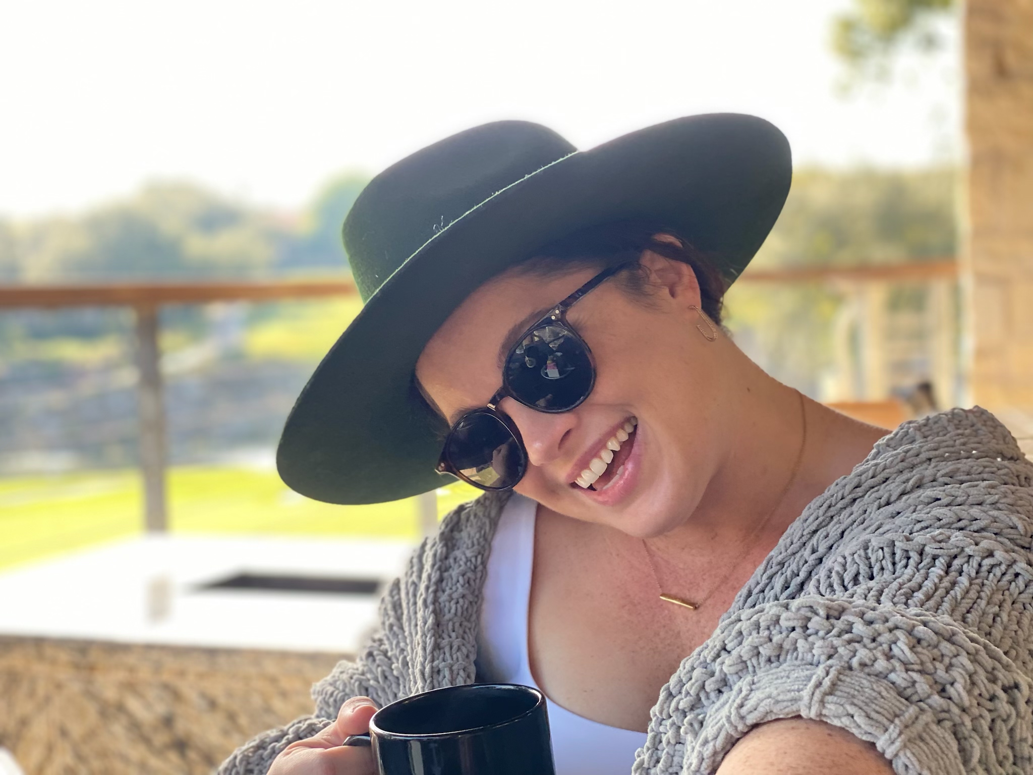 Woman wearing a large hat and sunglasses smiling outdoors, with a blurred background of trees and a fence.