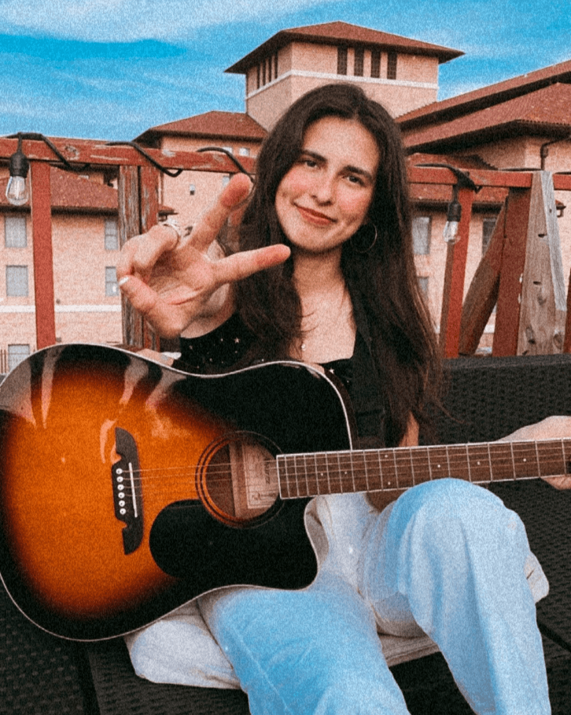 Young woman with long dark hair smiling, holding an acoustic guitar, making a peace sign outdoors with buildings in background.