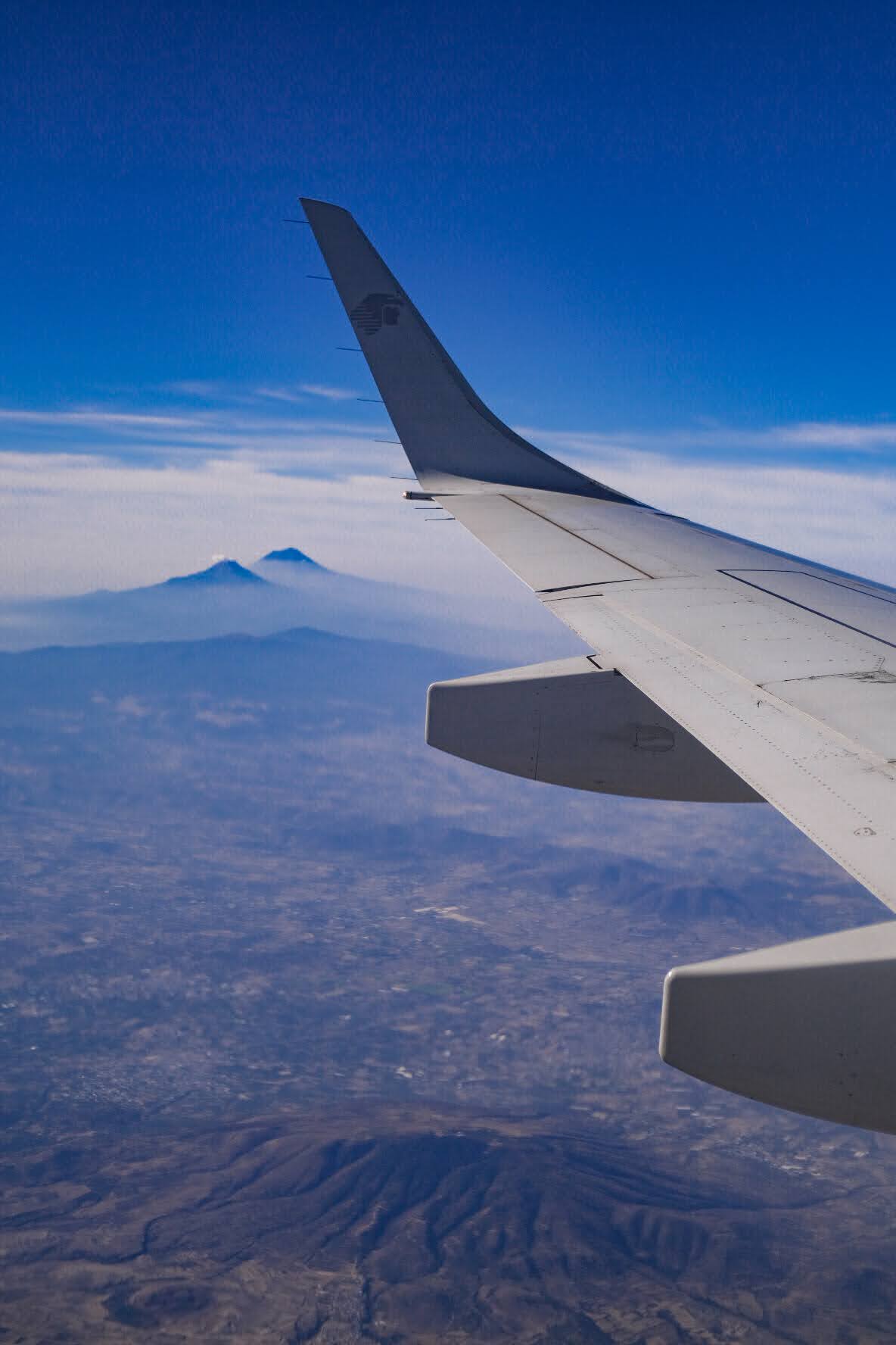View of an airplane wing with a mountain in the distance, above clouds, under a blue sky.
