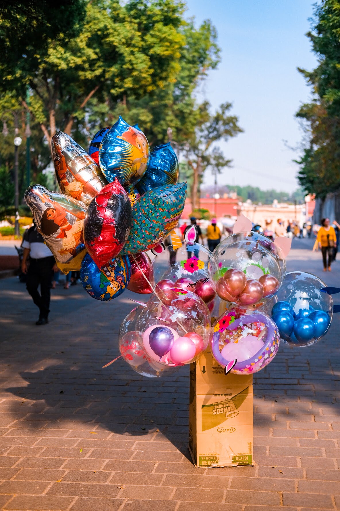 Balloon vendor's stand with colorful balloons on a sunny street, trees lining the sidewalk, and people walking in the background.