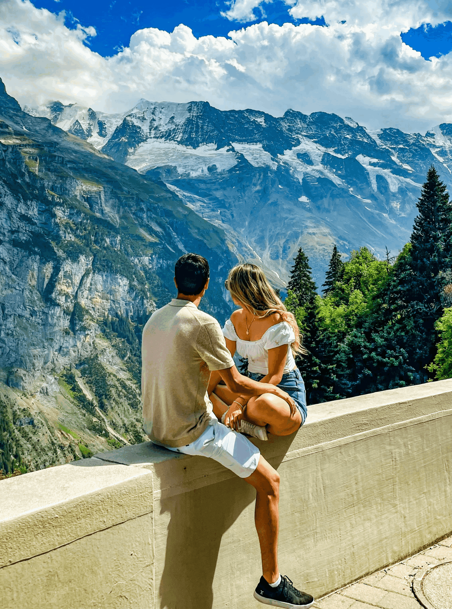 Two people sit on a ledge overlooking mountains and trees, with snow-capped peaks in the background, under a partly cloudy sky.