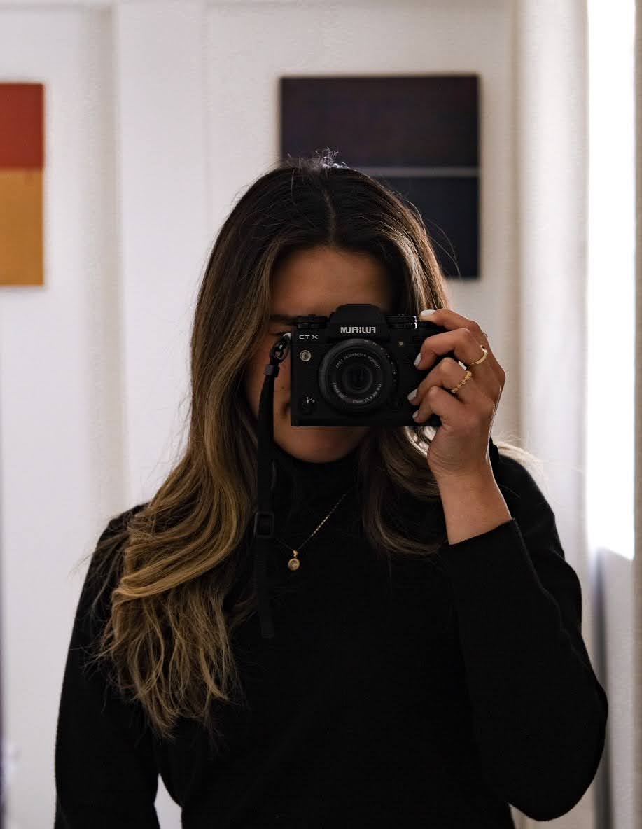Person with long hair holding a camera in front of their face, wearing a black top, indoors with artwork on the wall