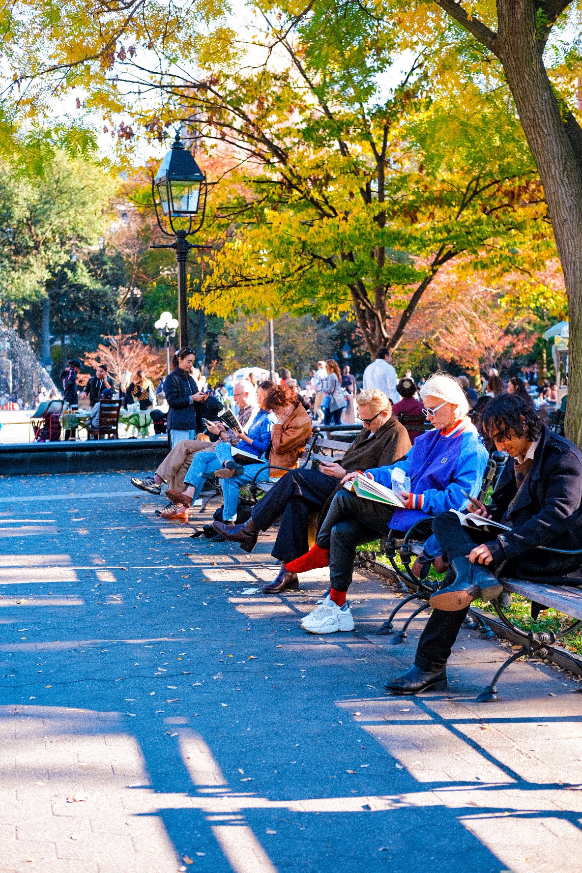People sitting on benches in a park with colorful autumn trees and a lamppost, sunny day.