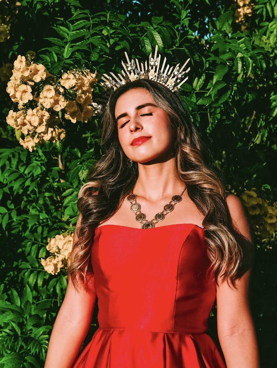 Young woman with long wavy hair wearing a red dress and a crown, standing outdoors with green foliage and flowers behind her.