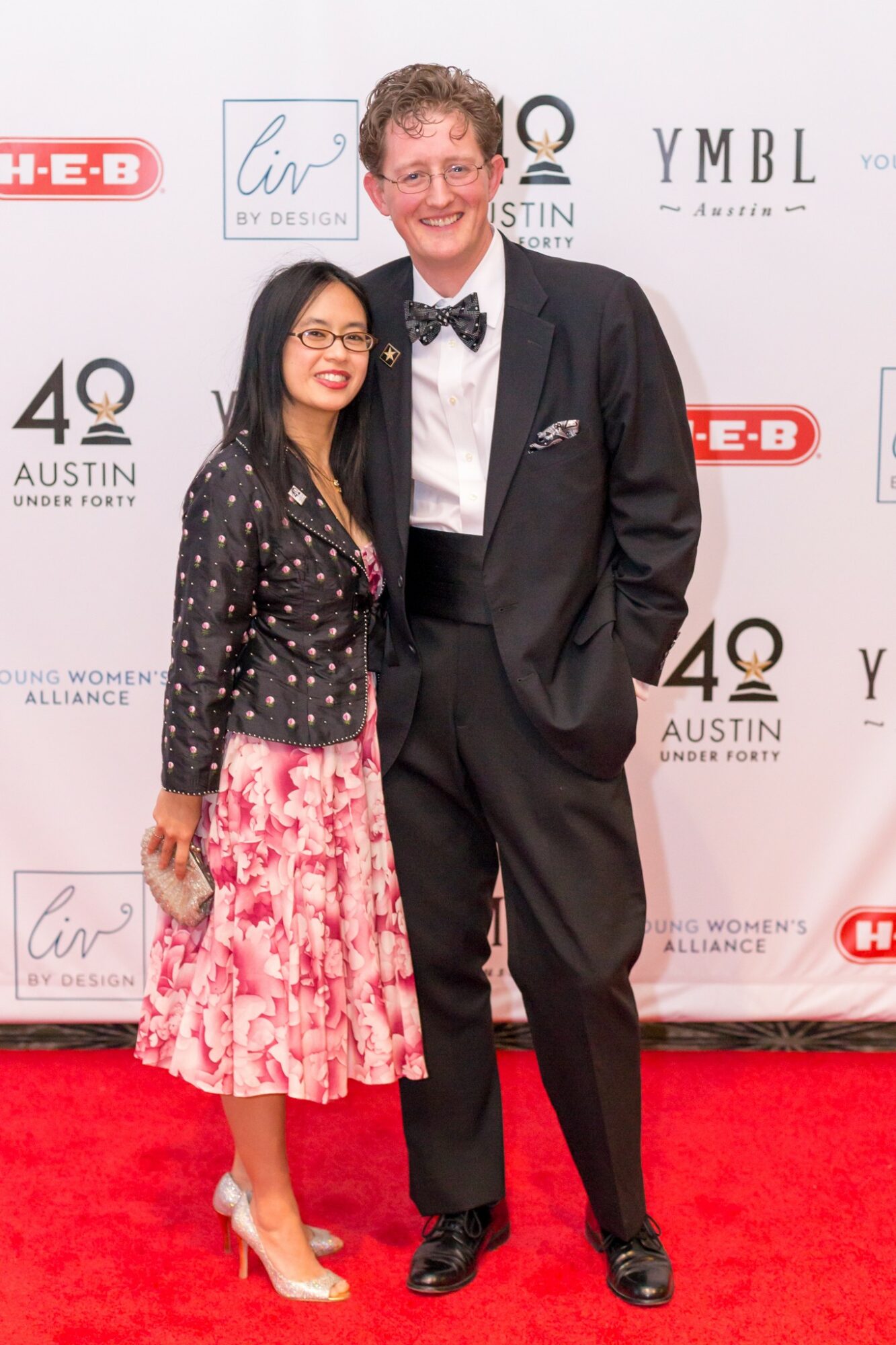 Two people standing on a red carpet, smiling, with event banners in the background.