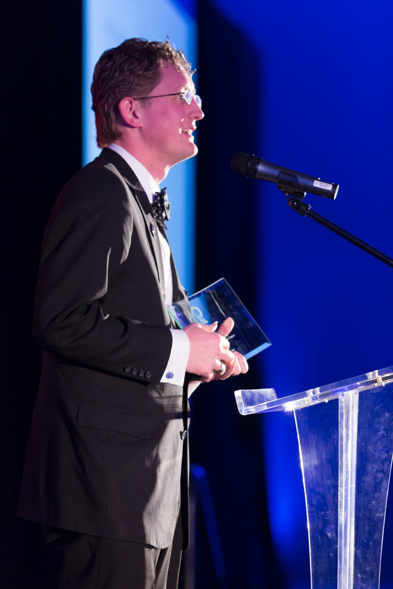 Man in tuxedo speaking at a podium with a microphone, holding a glass award, against a blue background.