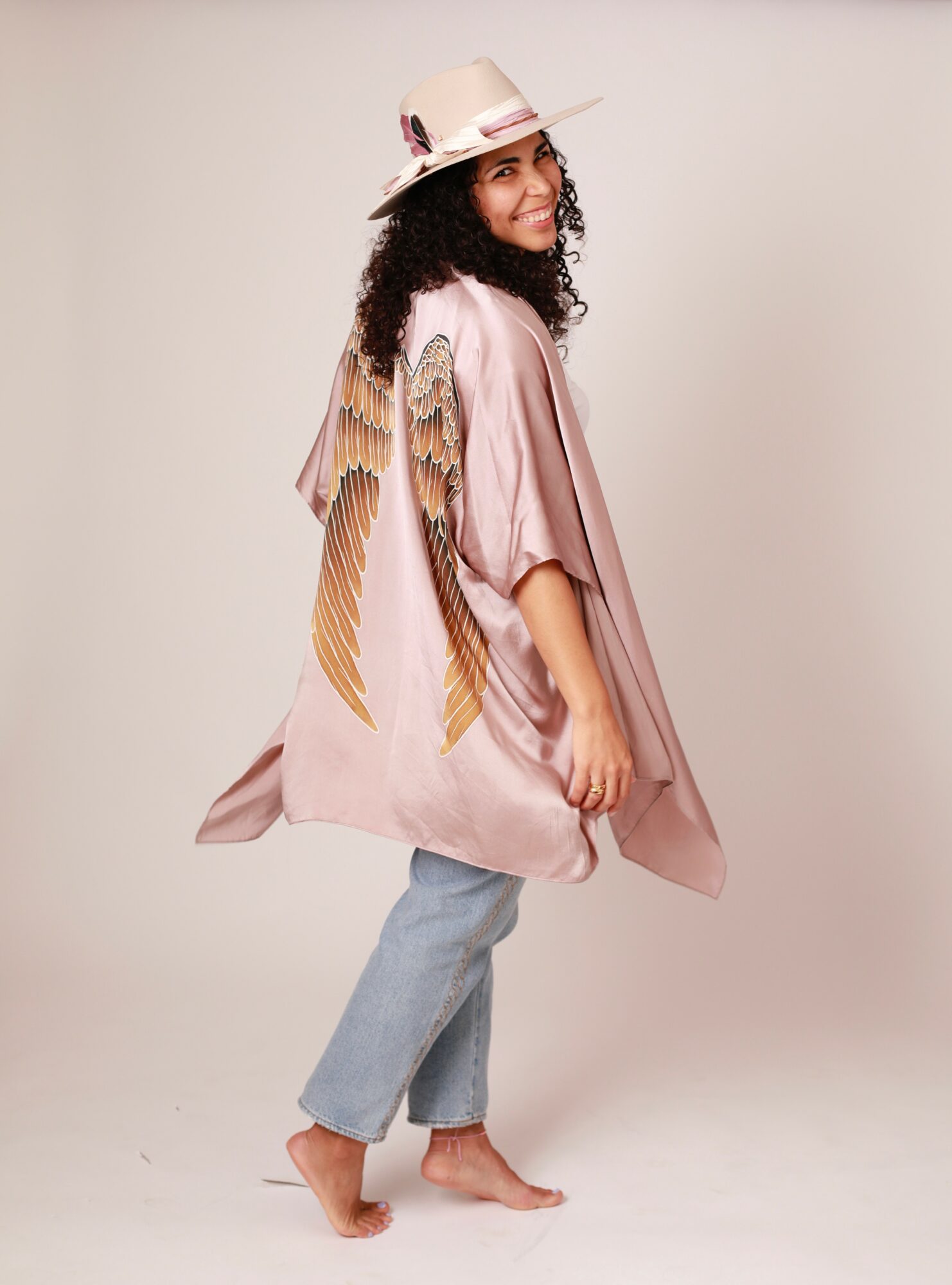 Woman with curly hair wearing a hat, pink cape with wings, and jeans, standing barefoot against a plain background.