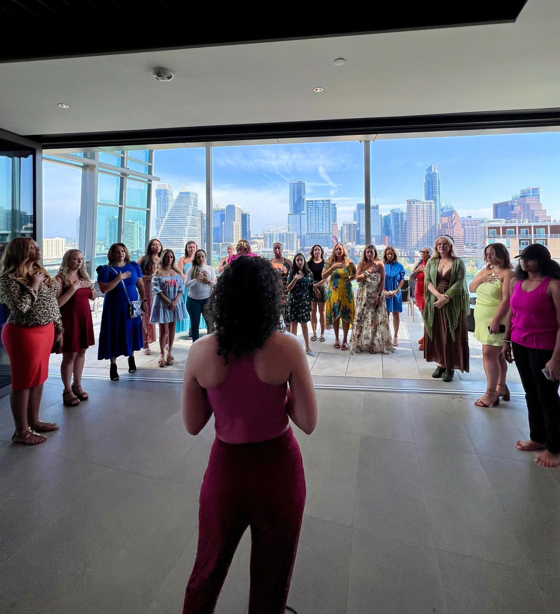 Group of women standing in a circle indoors with a city skyline view outside large windows, woman in pink facing away.