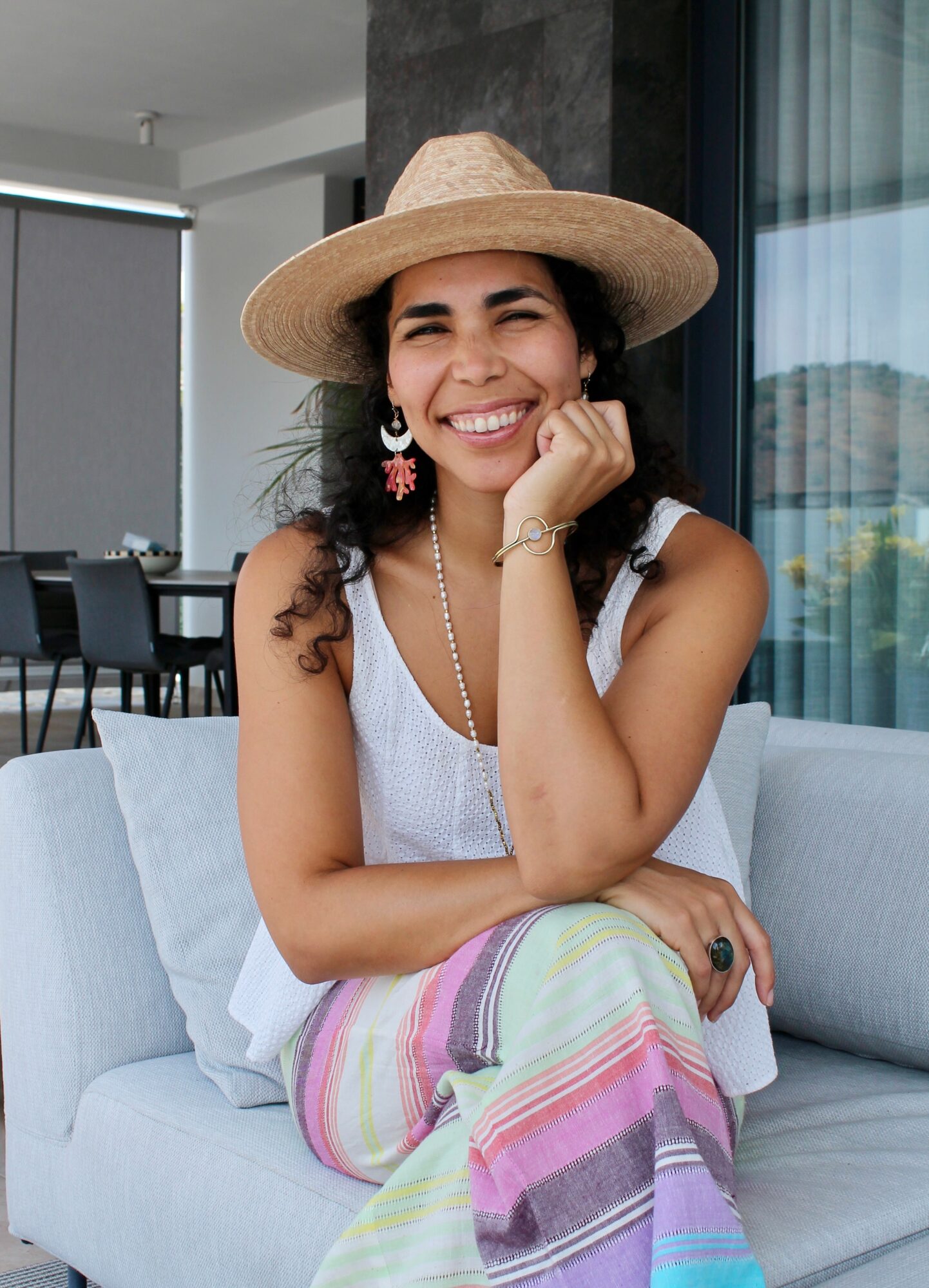 Woman smiling, wearing a straw hat, white top, and colorful striped pants, sitting on a sofa indoors.