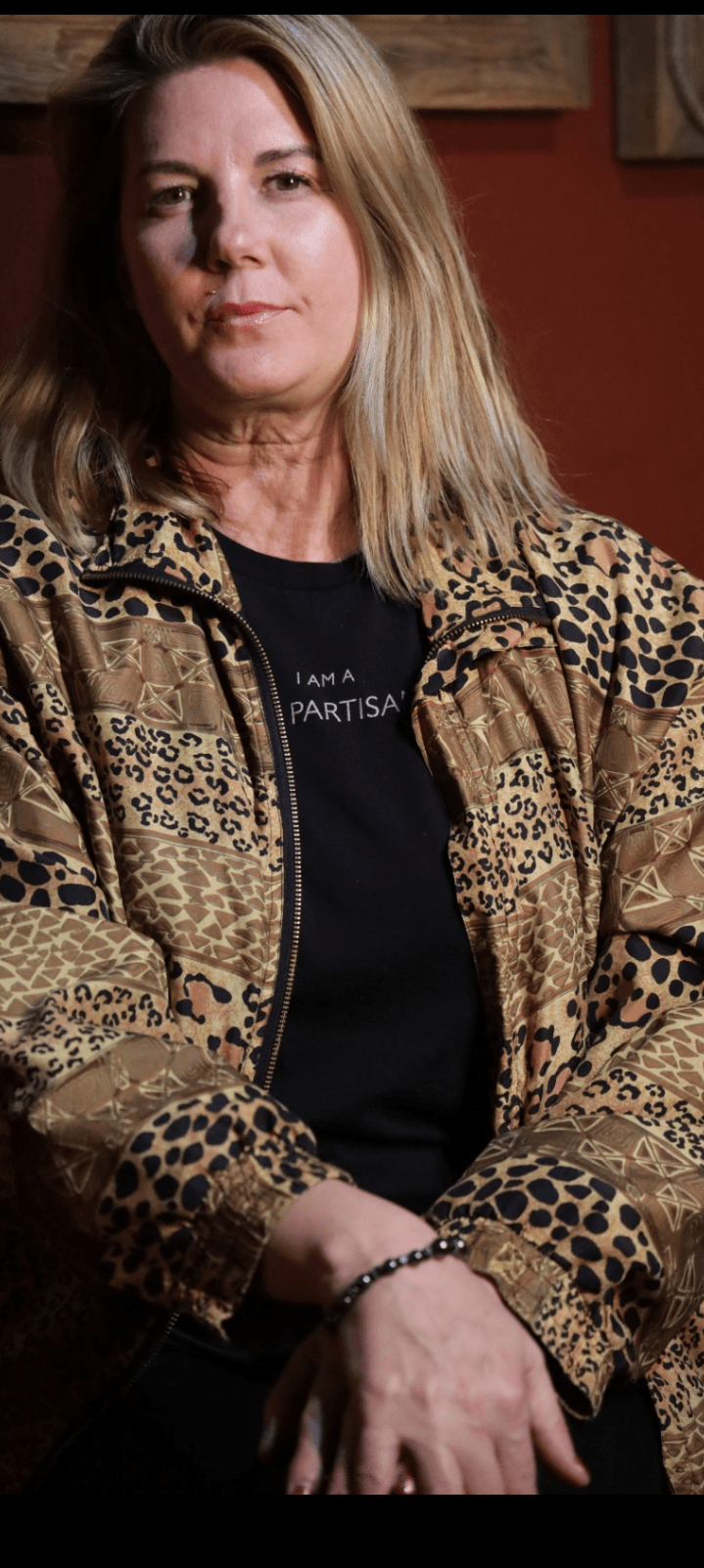 Woman with shoulder-length hair wearing a patterned jacket and black shirt, sitting with hands crossed.