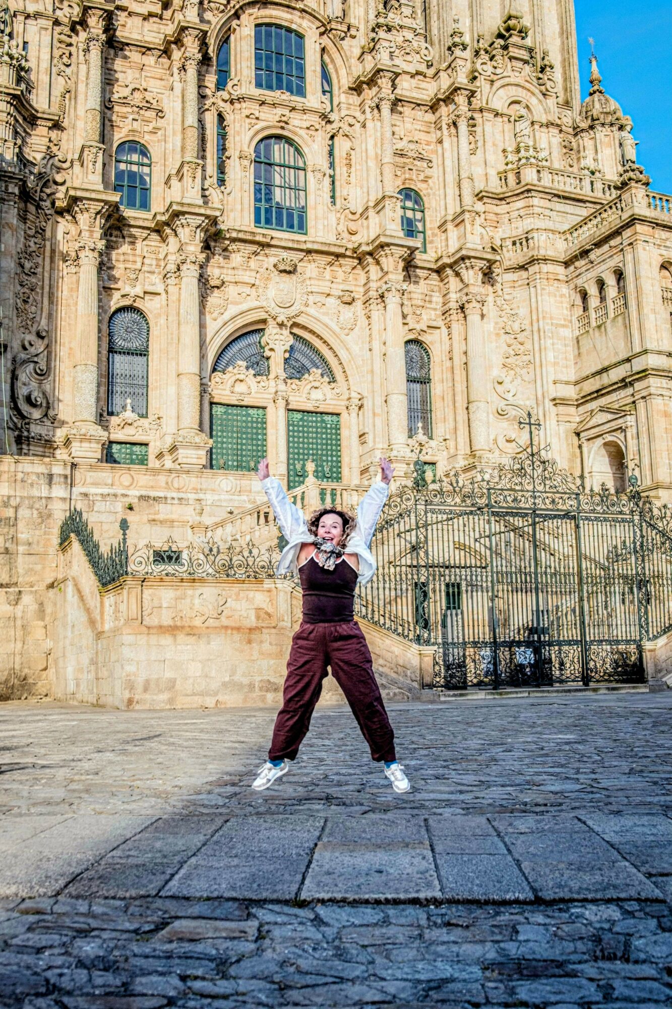 Person standing with arms raised in front of an ornate historic building, wearing a hat and casual clothing.
