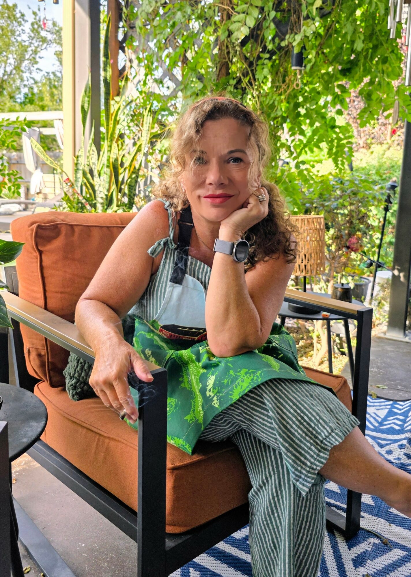 Woman sitting on a sofa outdoors, resting her chin on her hand, surrounded by greenery and sunlight.