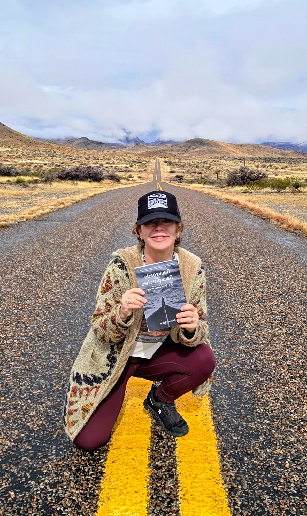 Person crouching on a road holding a book, landscape with hills and cloudy sky in background.