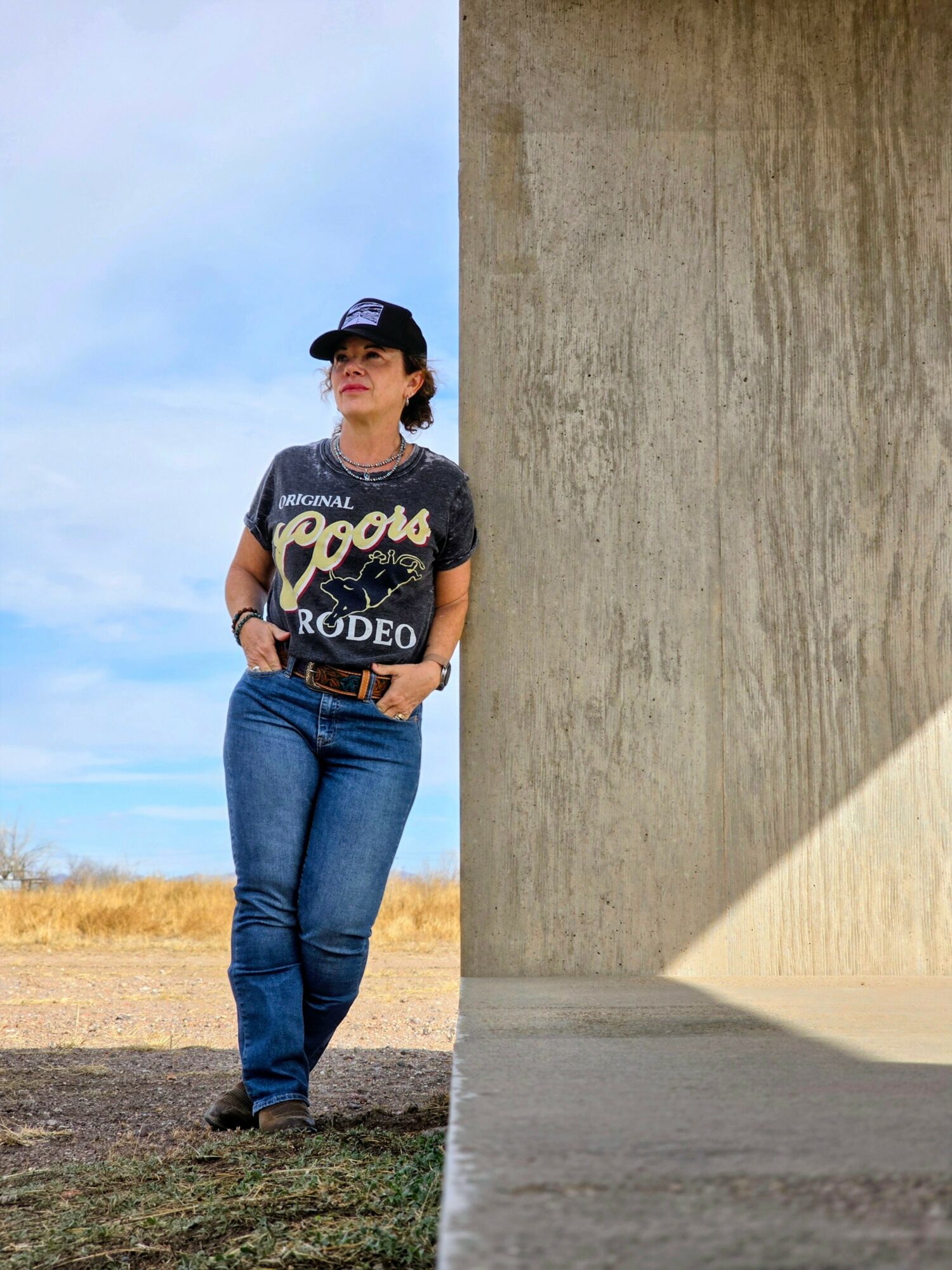 Woman standing outdoors next to a large concrete wall, wearing a cap, t-shirt, and jeans, with a blue sky background.