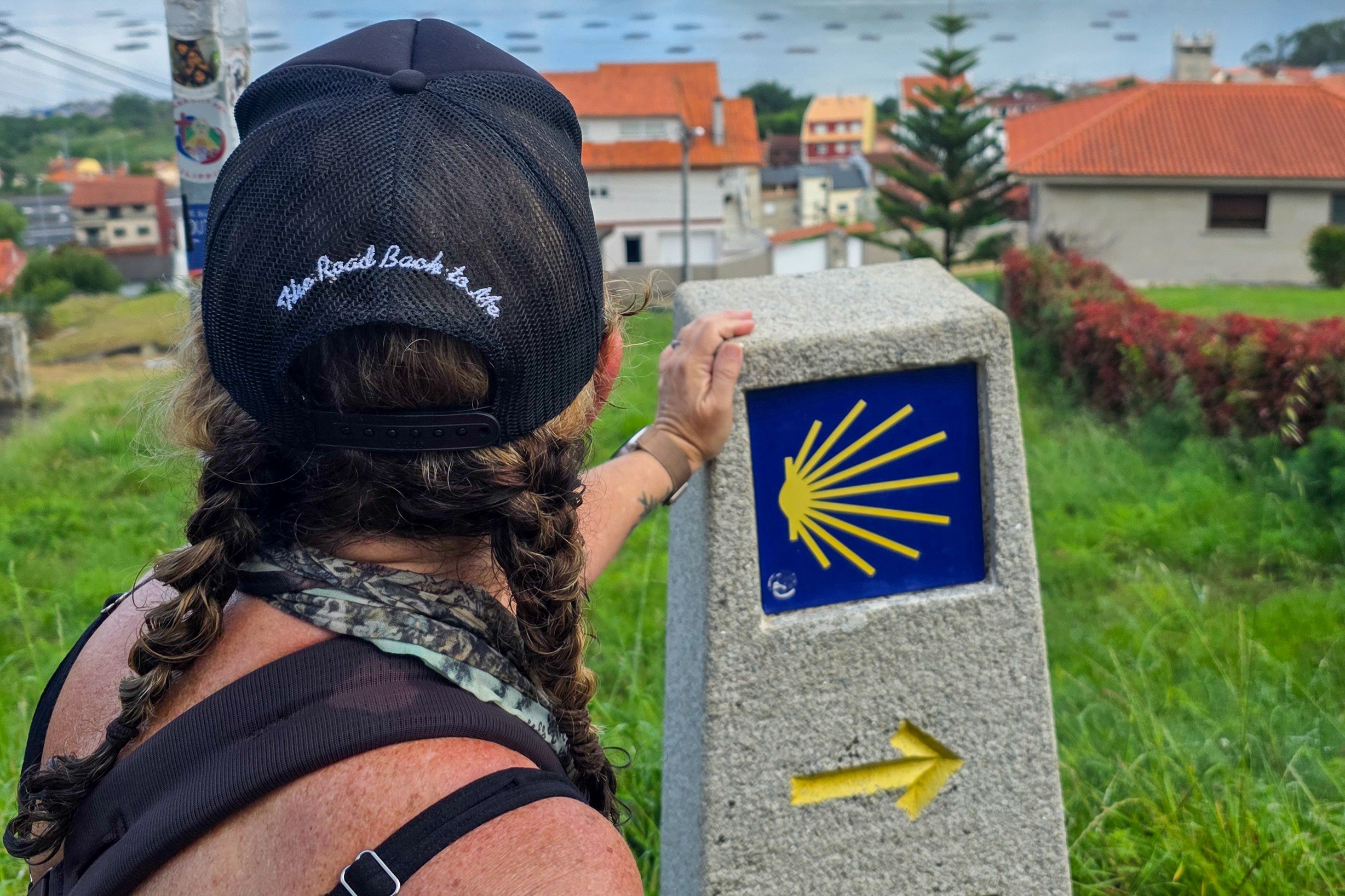 Child with braided hair and a cap touching a stone marker with a yellow arrow and a yellow scallop shell symbol, outdoors.