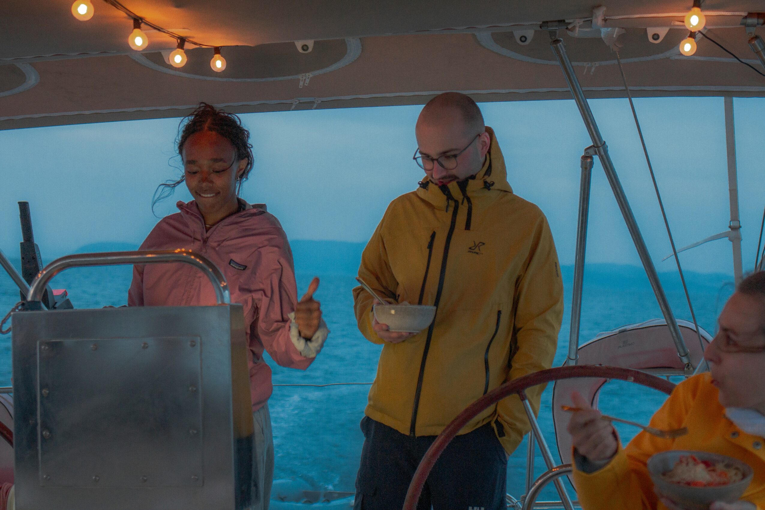 Two people on a boat, one holding a bowl, with ocean and sky in background, under string lights.