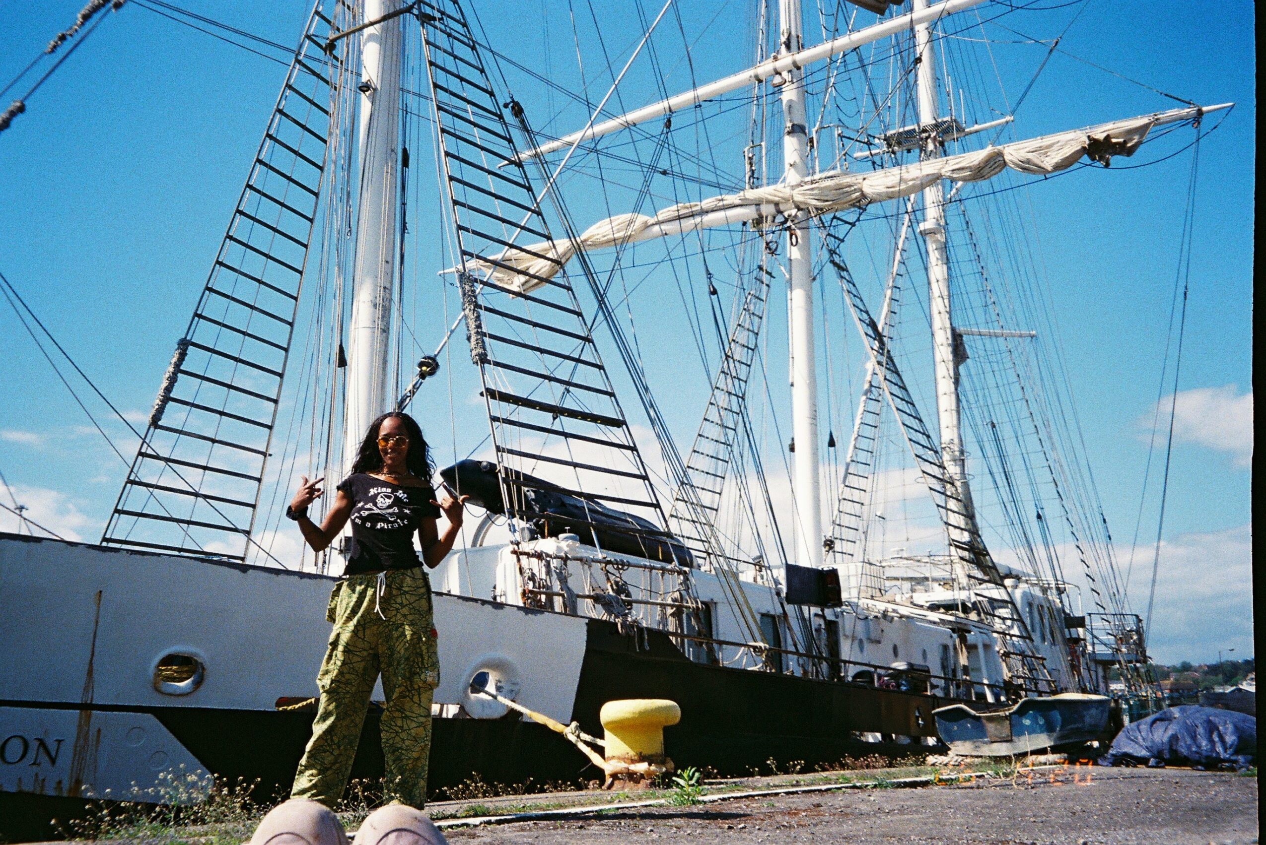 Person standing on dock near large sailing ship with tall masts and rigging, blue sky background.