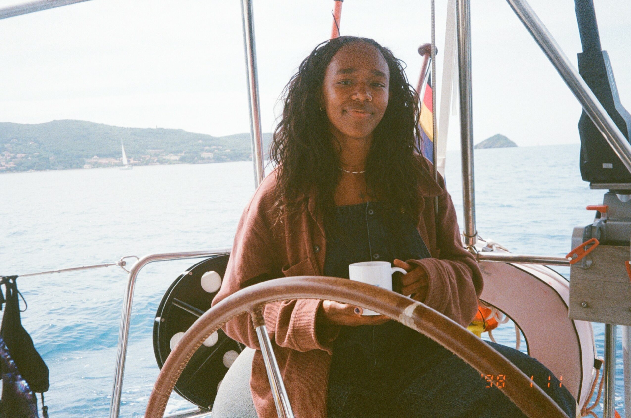 Person with long hair standing on a boat holding a mug, with water and land in the background.