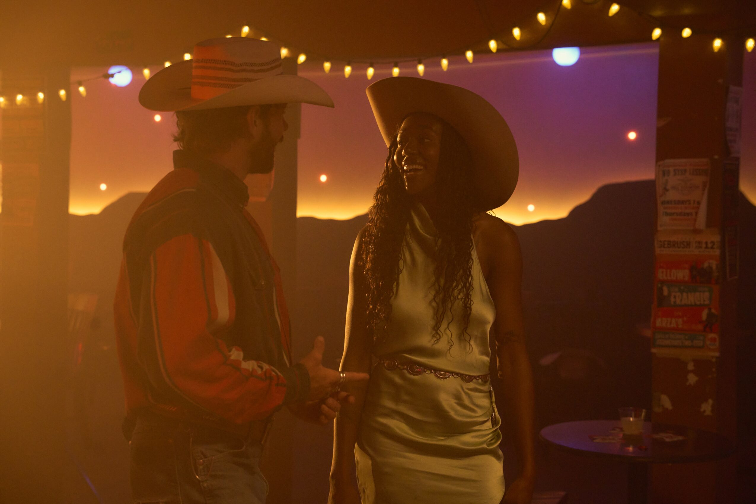 Two people wearing hats and casual clothing stand in a dimly lit setting with string lights and a sunset background.