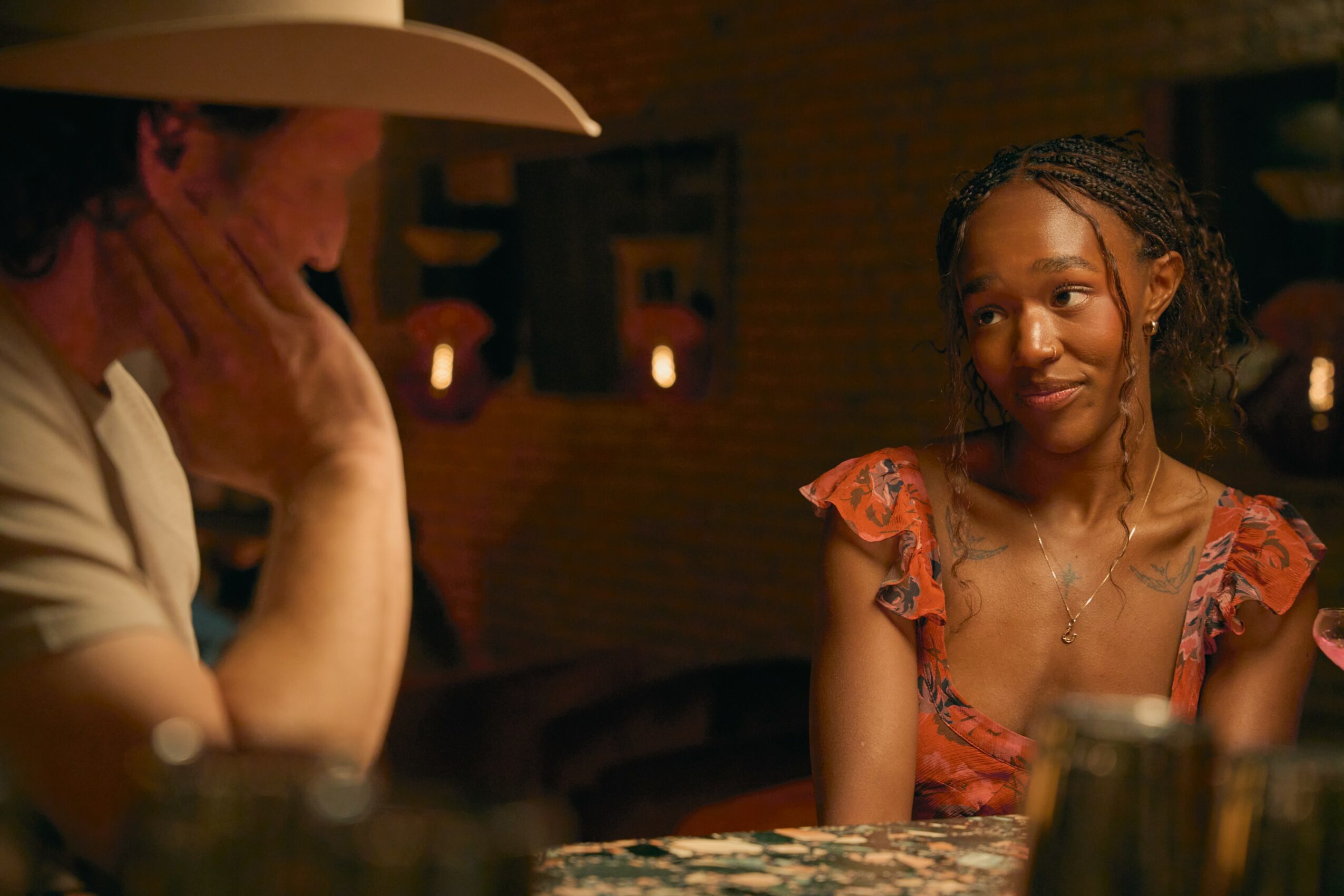 Two women sitting at a table in a dimly lit setting, one wearing a hat and the other in a floral dress, smiling.