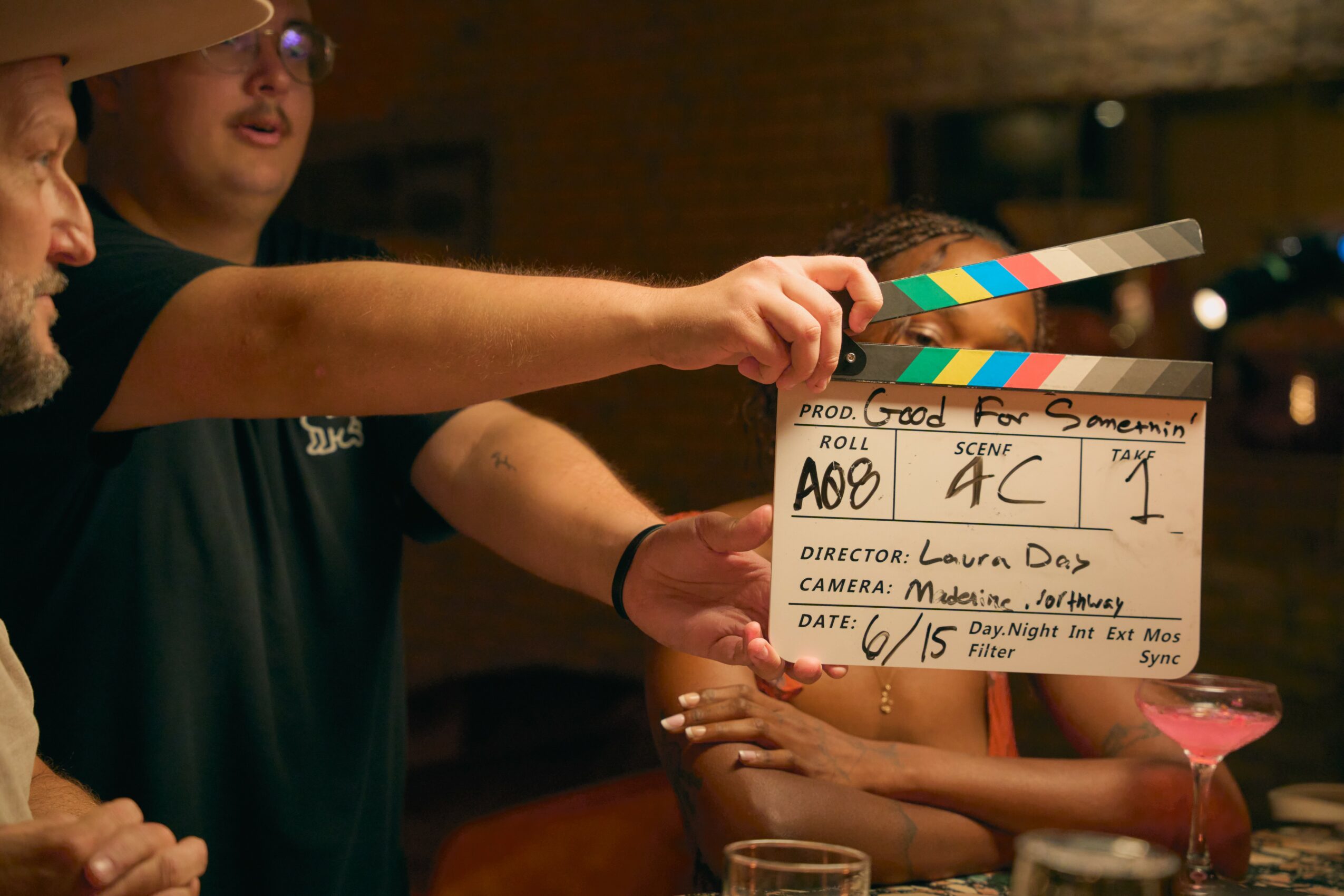 Two people hold a clapperboard on a film set, with a glass of pink drink visible in the foreground.