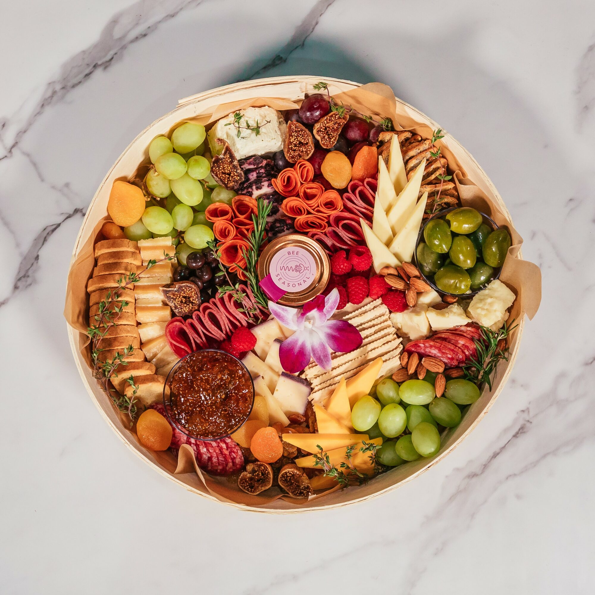 Round platter with grapes, cheese, crackers, meats, and fruits on a marble surface.