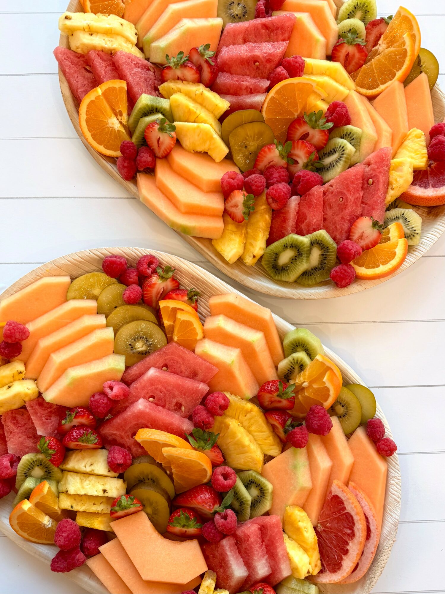 Two plates of assorted sliced fruits including watermelon, cantaloupe, kiwi, pineapple, oranges, and berries on a white surface.