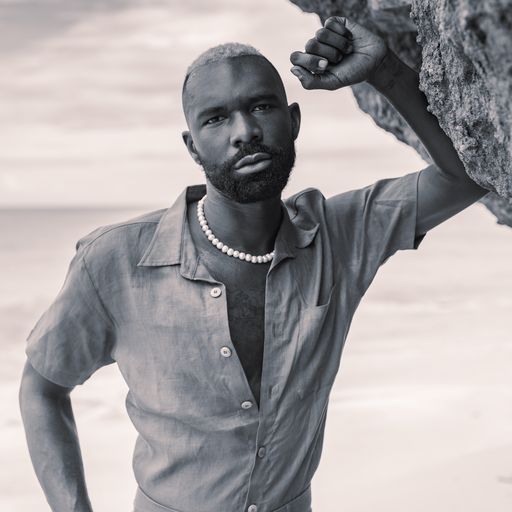 Man with a beard and pearl necklace leaning on a rock, wearing a button-up shirt, outdoors, black and white photo.