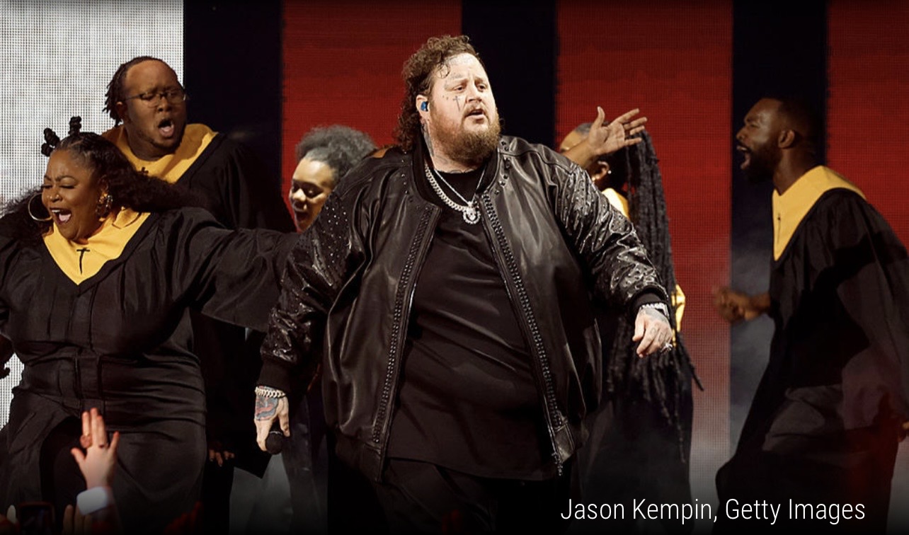 Group of people singing on stage, one person in the center wearing a black leather jacket, others in black and yellow robes.