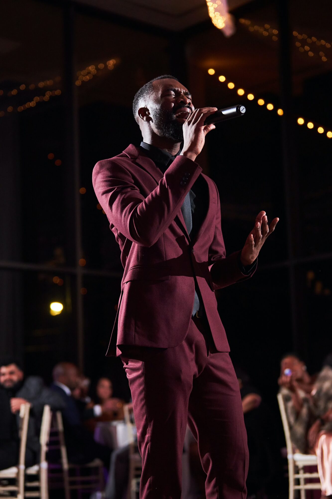 Man in burgundy suit singing into a microphone at an indoor event with seated audience.