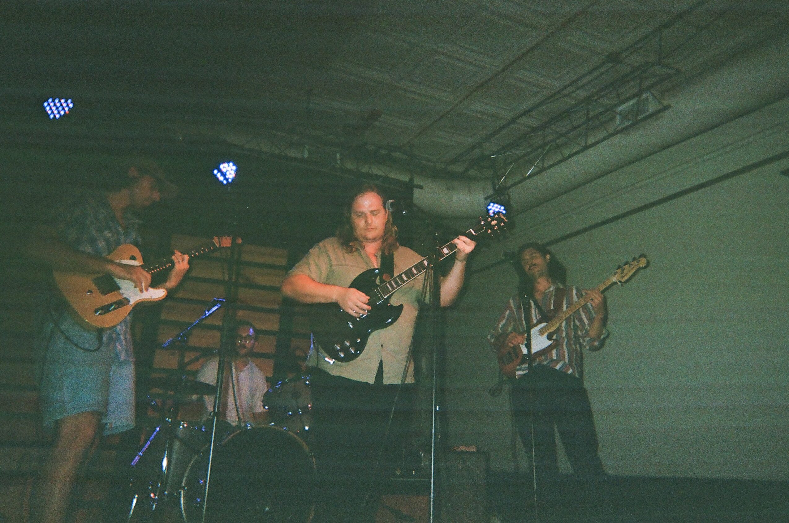 Three musicians perform on stage with guitars, under stage lights, in a dimly lit setting.