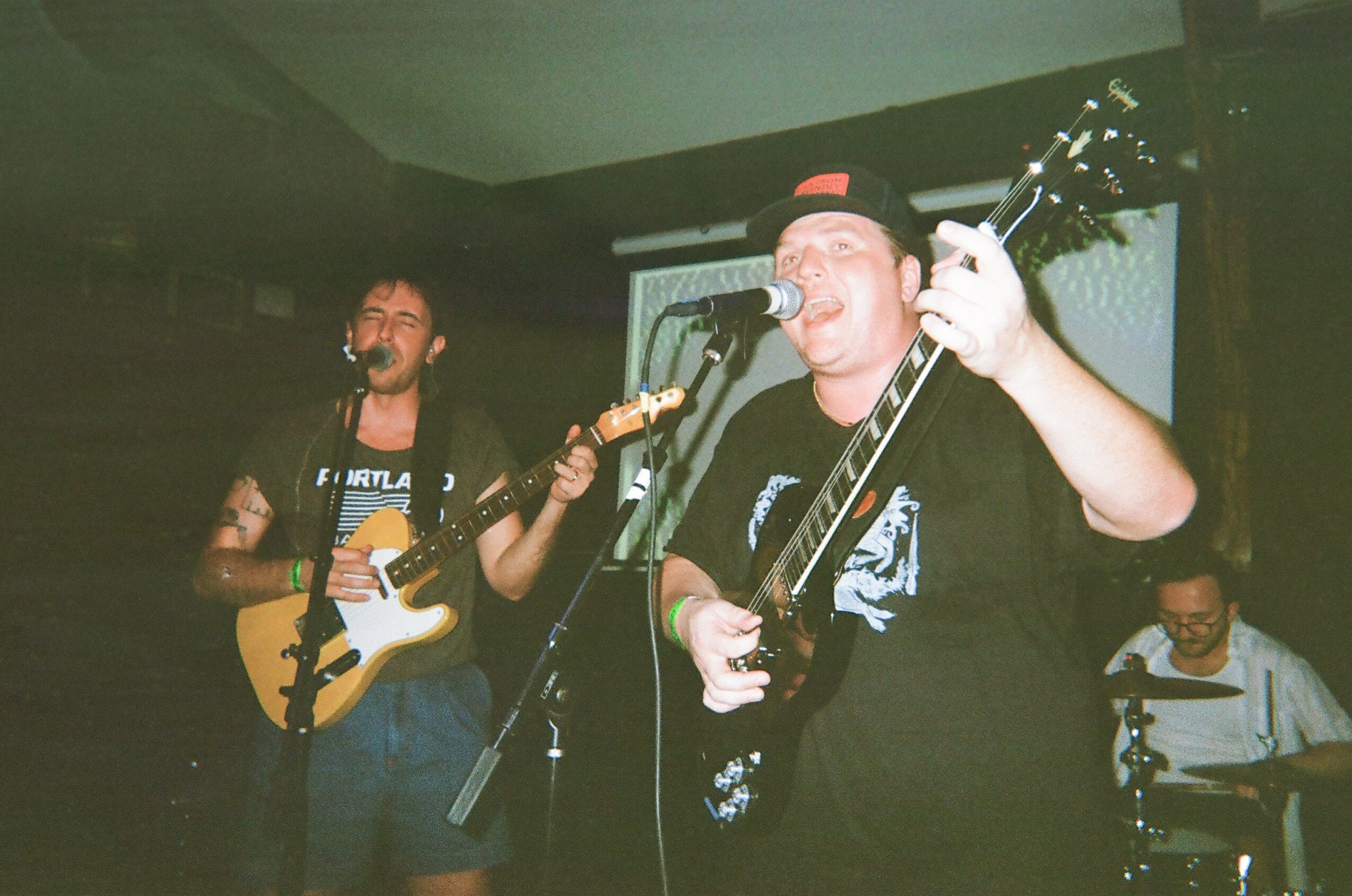 Three musicians perform on stage, two playing guitars and singing, one on drums, in a dimly lit setting.
