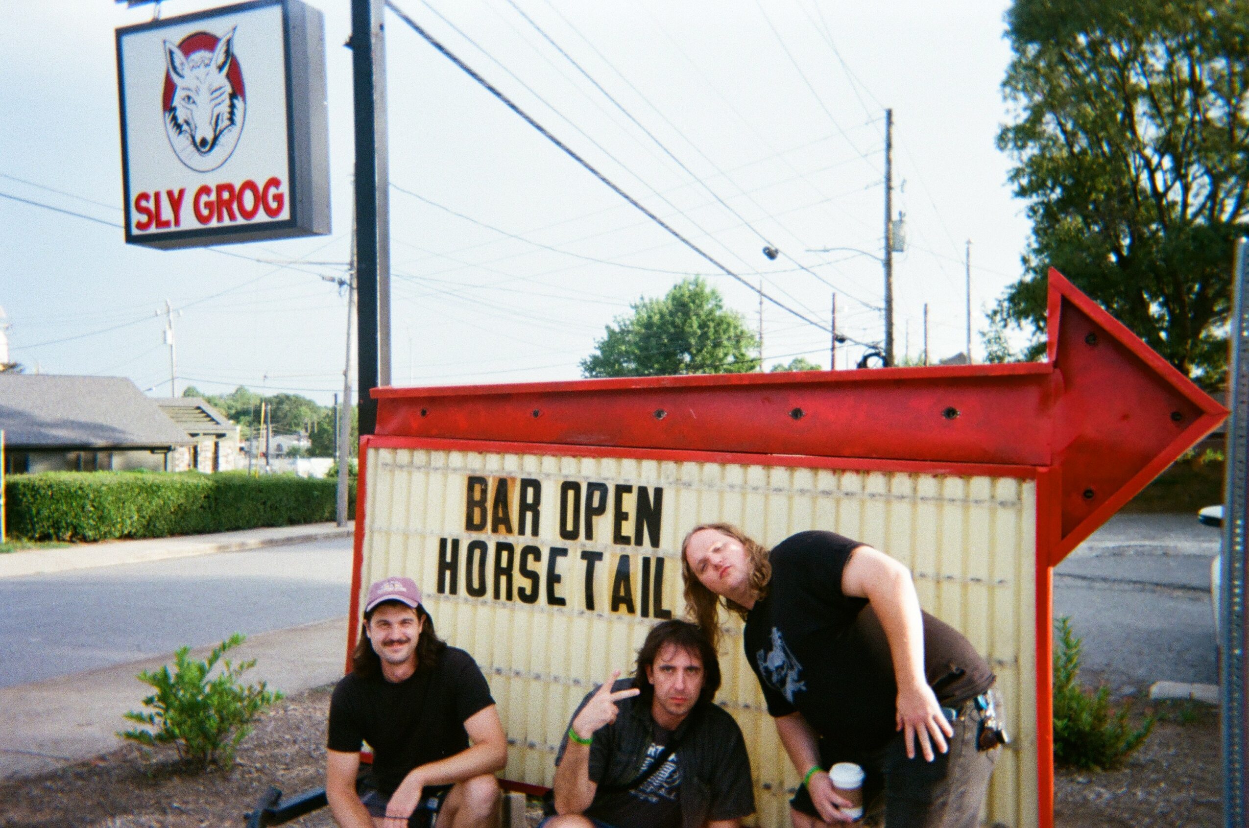Three people sitting and leaning in front of a sign that reads 'Bar Open Horse Tails' with a red arrow pointing right, outside a business with a sign that says 'Sly Grog'.