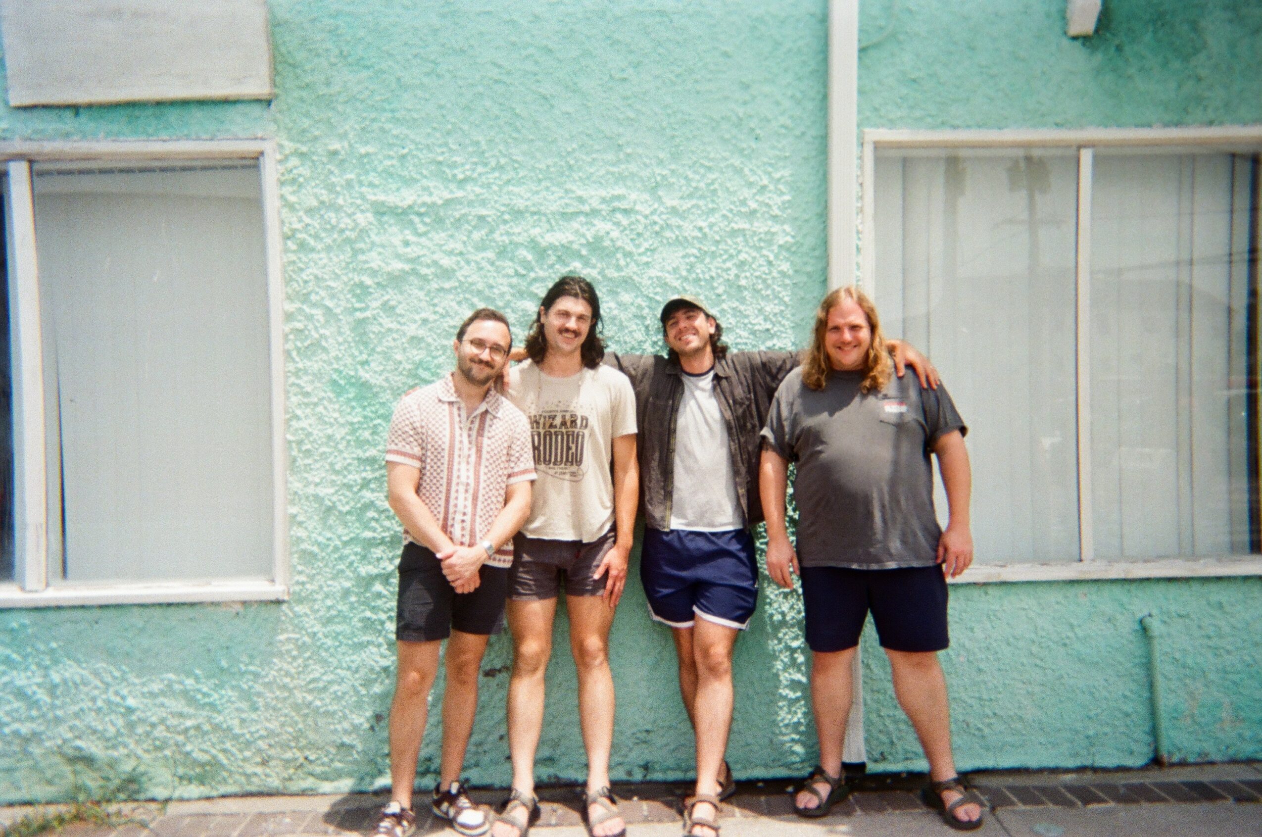 Four people standing in front of a turquoise wall with windows, smiling and posing together.