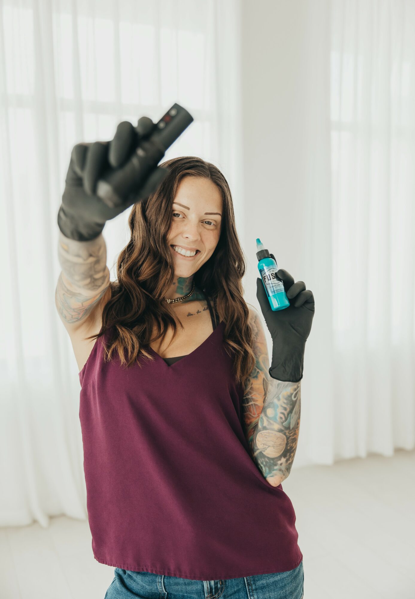 Young woman with long hair, wearing a purple top and black gloves, holding a tattoo machine and ink bottle, smiling.