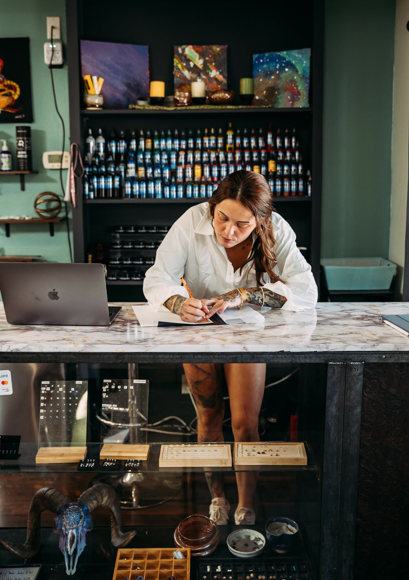 Woman in white shirt at bar counter, looking at her phone, with laptop and bottles behind her, colorful artwork above.