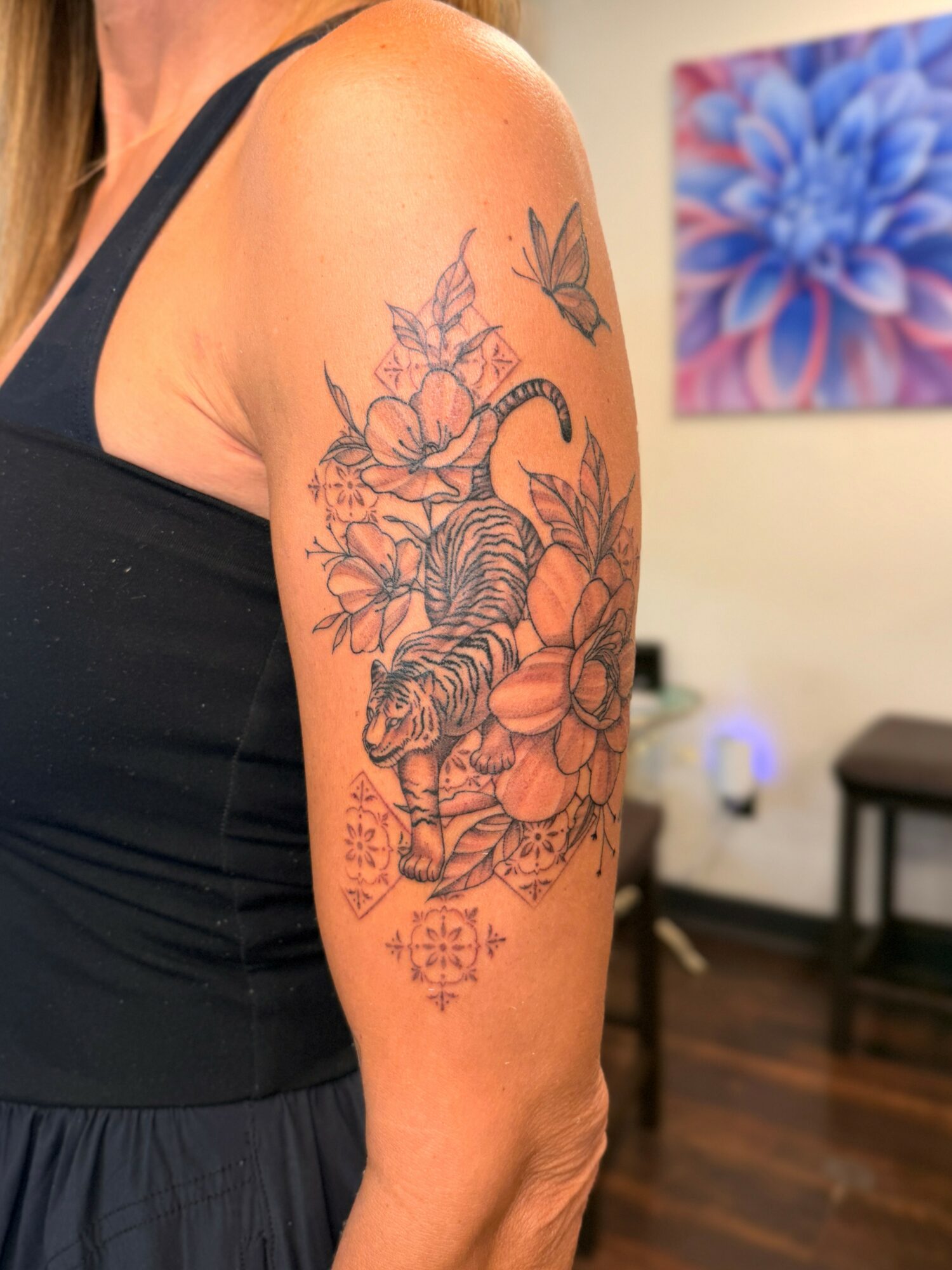 Tattoo of a tiger surrounded by flowers and a butterfly on person's upper arm, with a colorful flower painting in background.
