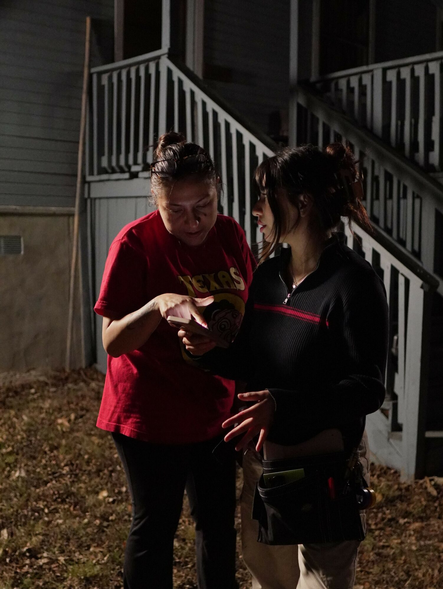 Two women standing outdoors near a staircase, one in a red shirt and the other in black, engaged in conversation.