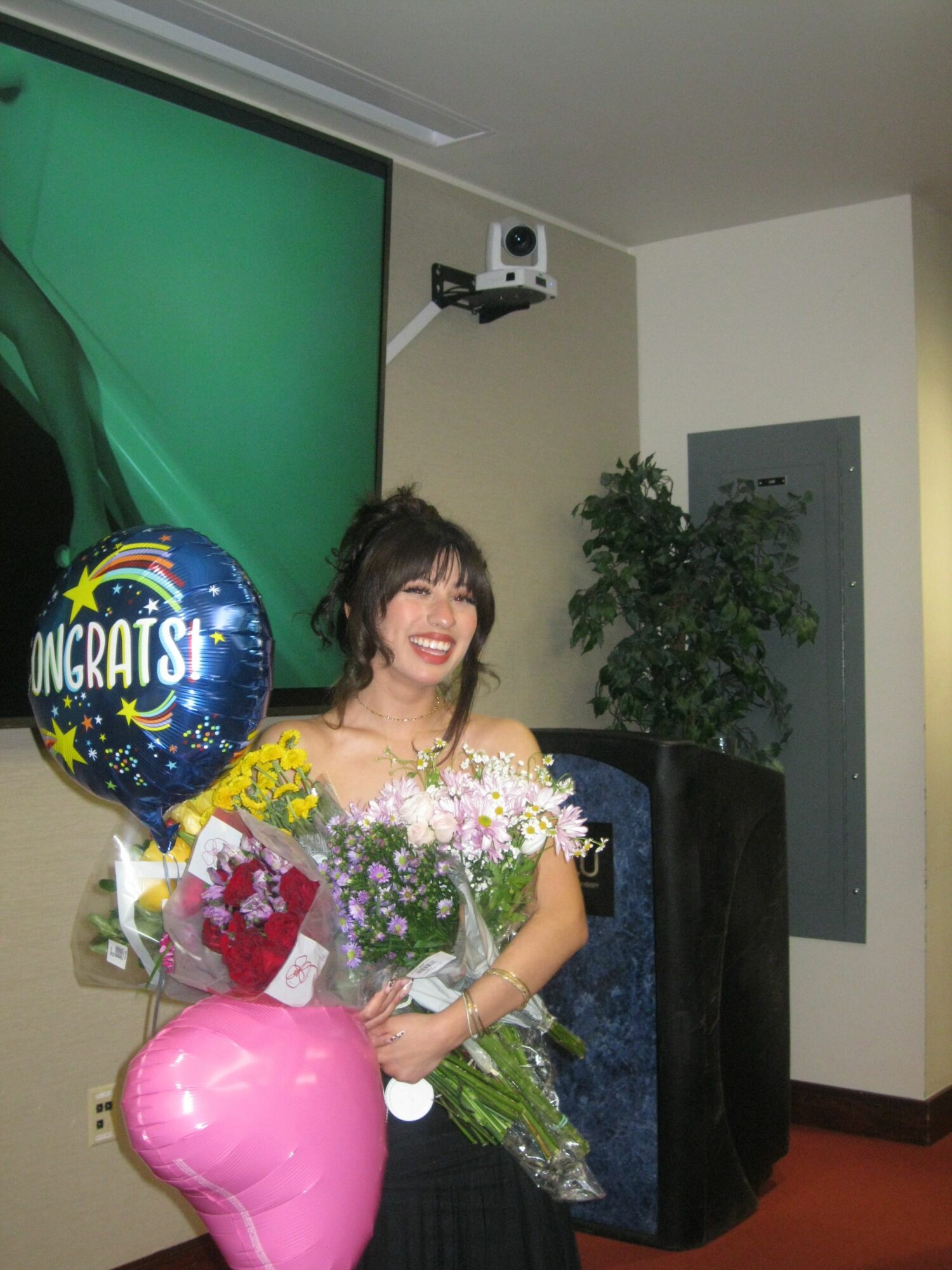 Woman holding flowers and balloons, smiling, in a room with a green screen and a potted plant.