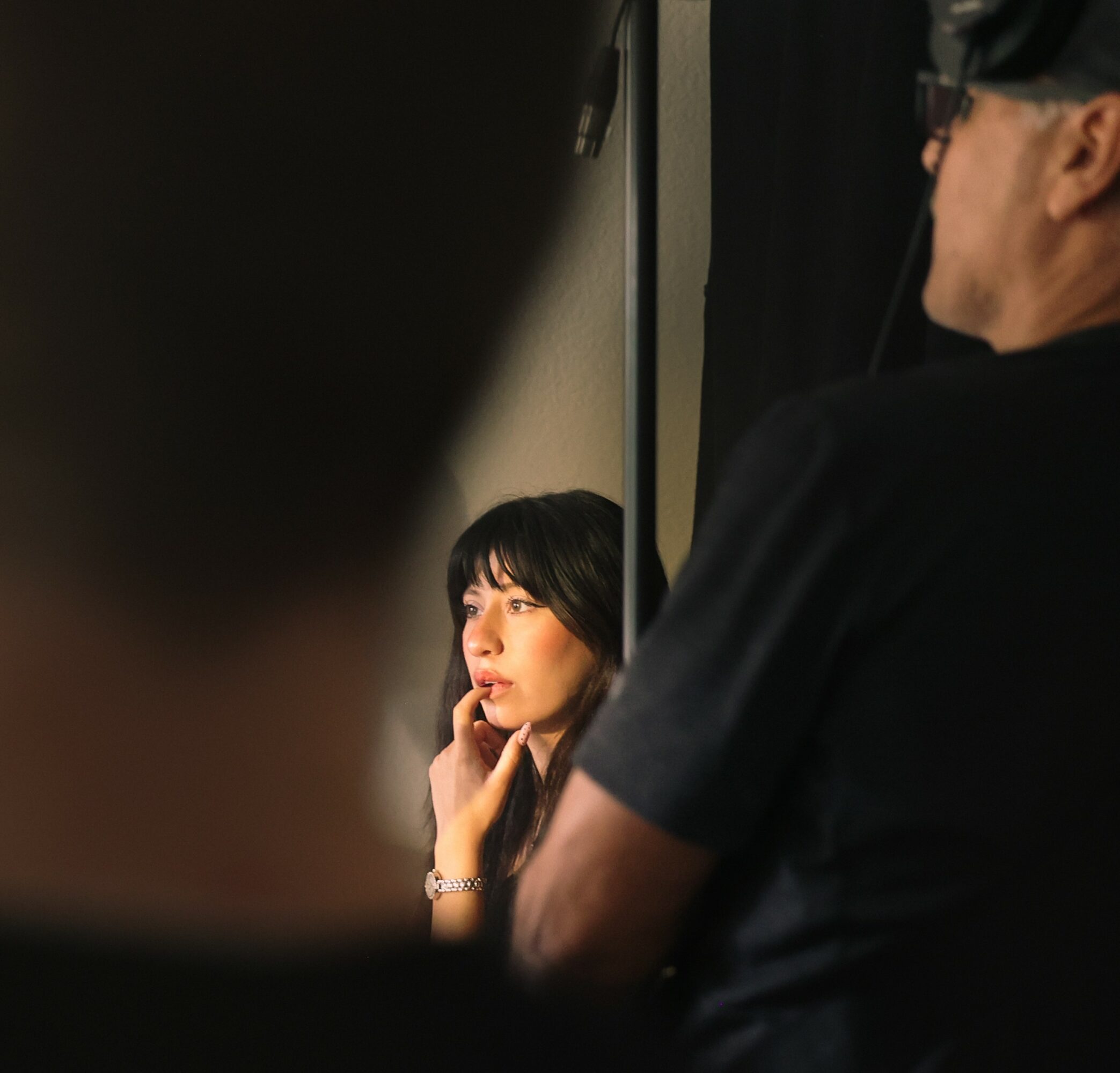 Woman with dark hair and makeup, resting her chin on her hand, looking to the side, with a person in black in the foreground.