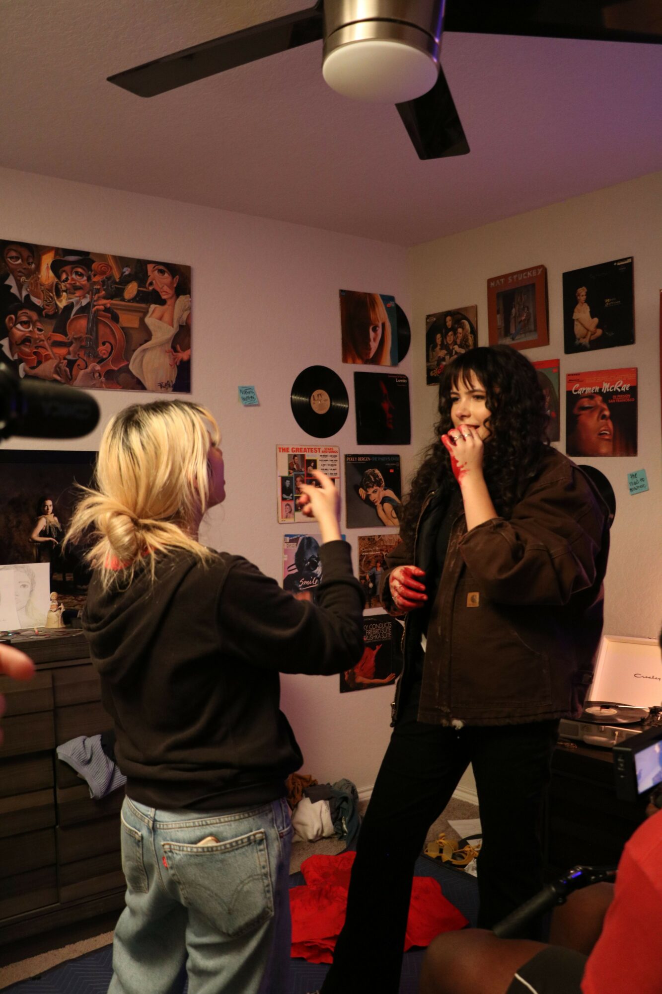 Two people with long hair, one with blonde and one with dark hair, are looking at photos on a wall in a room.