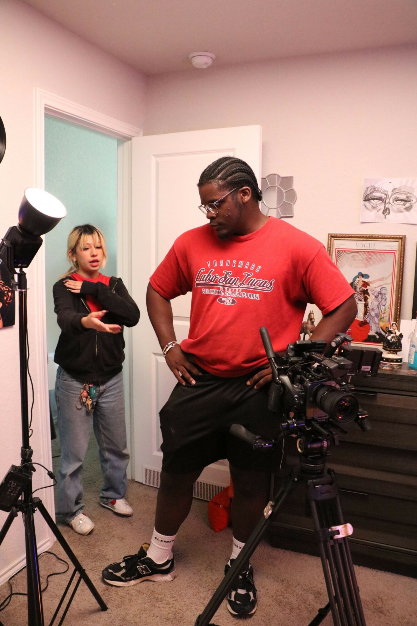 A woman in a red shirt and glasses stands behind a camera setup, with a woman in a black jacket and gray pants in the background, in a room with pink walls.