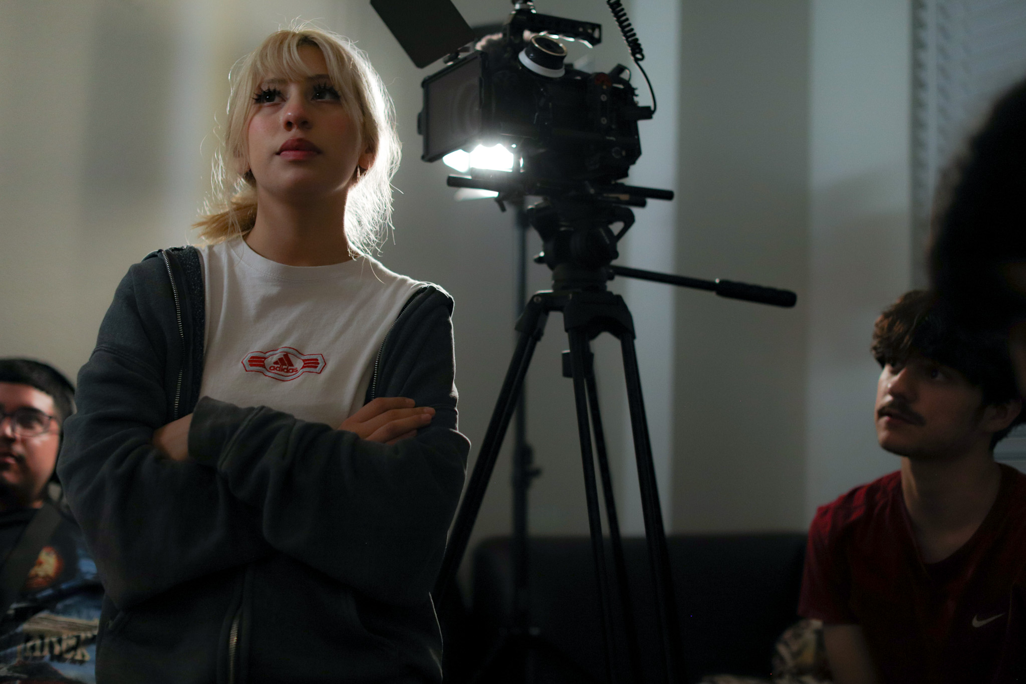 Young woman with blonde hair standing near a camera on a tripod, in a room with white walls and people around.