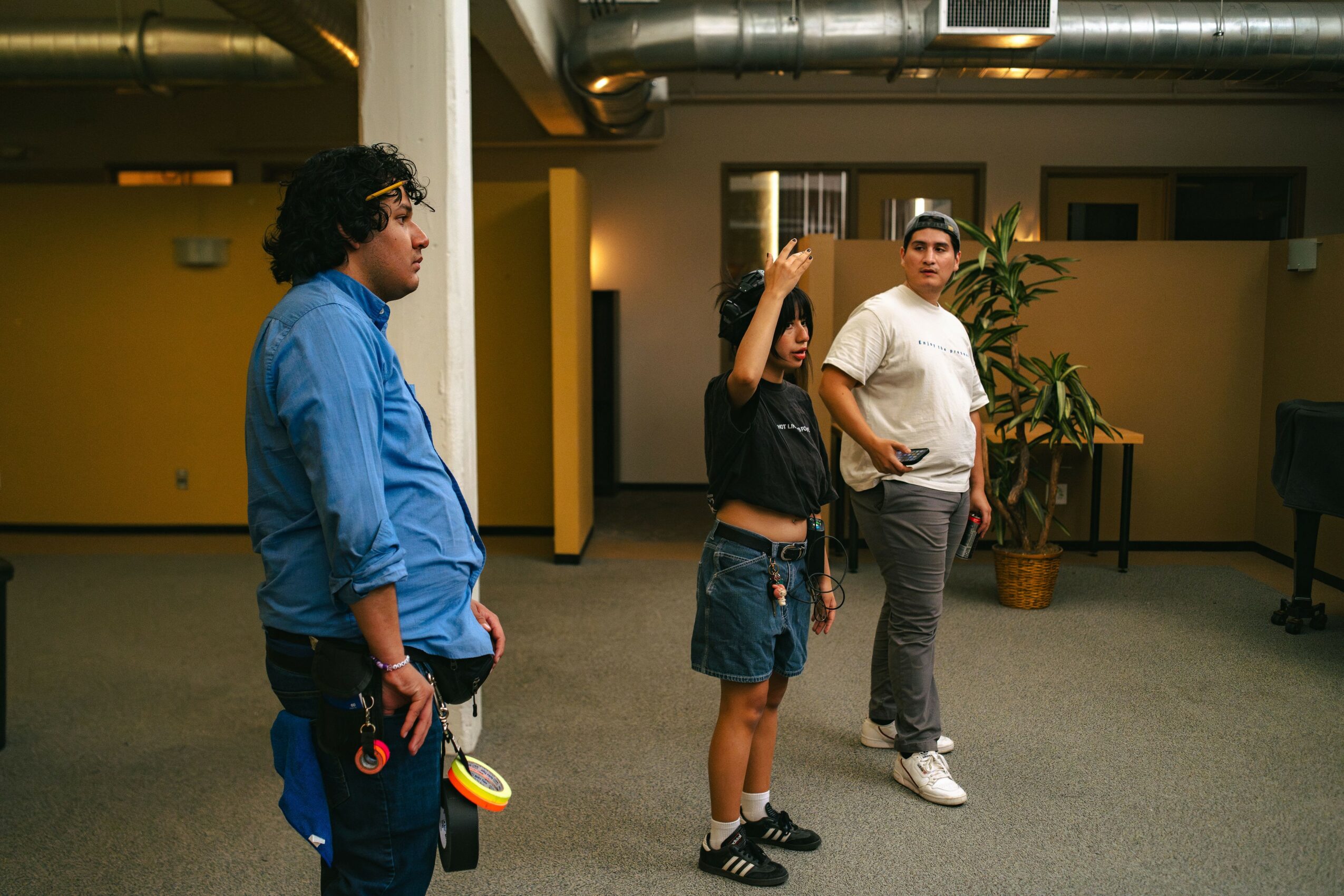 Three people standing and talking in an indoor space with yellow walls and plants, two facing each other and one looking away.