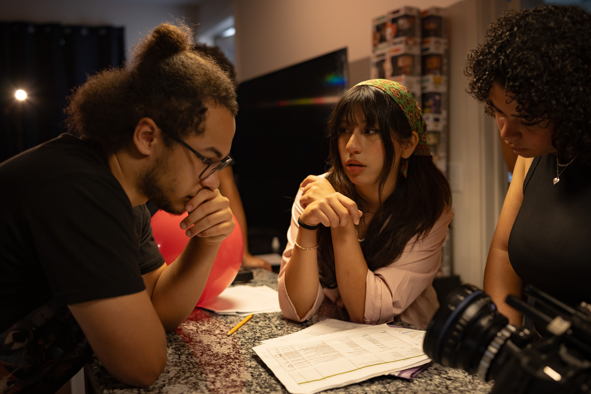 Three people sit around a table with papers, engaged in conversation, in a dimly lit room. One person has glasses and curly hair, another has long dark hair, and the third has short curly hair.
