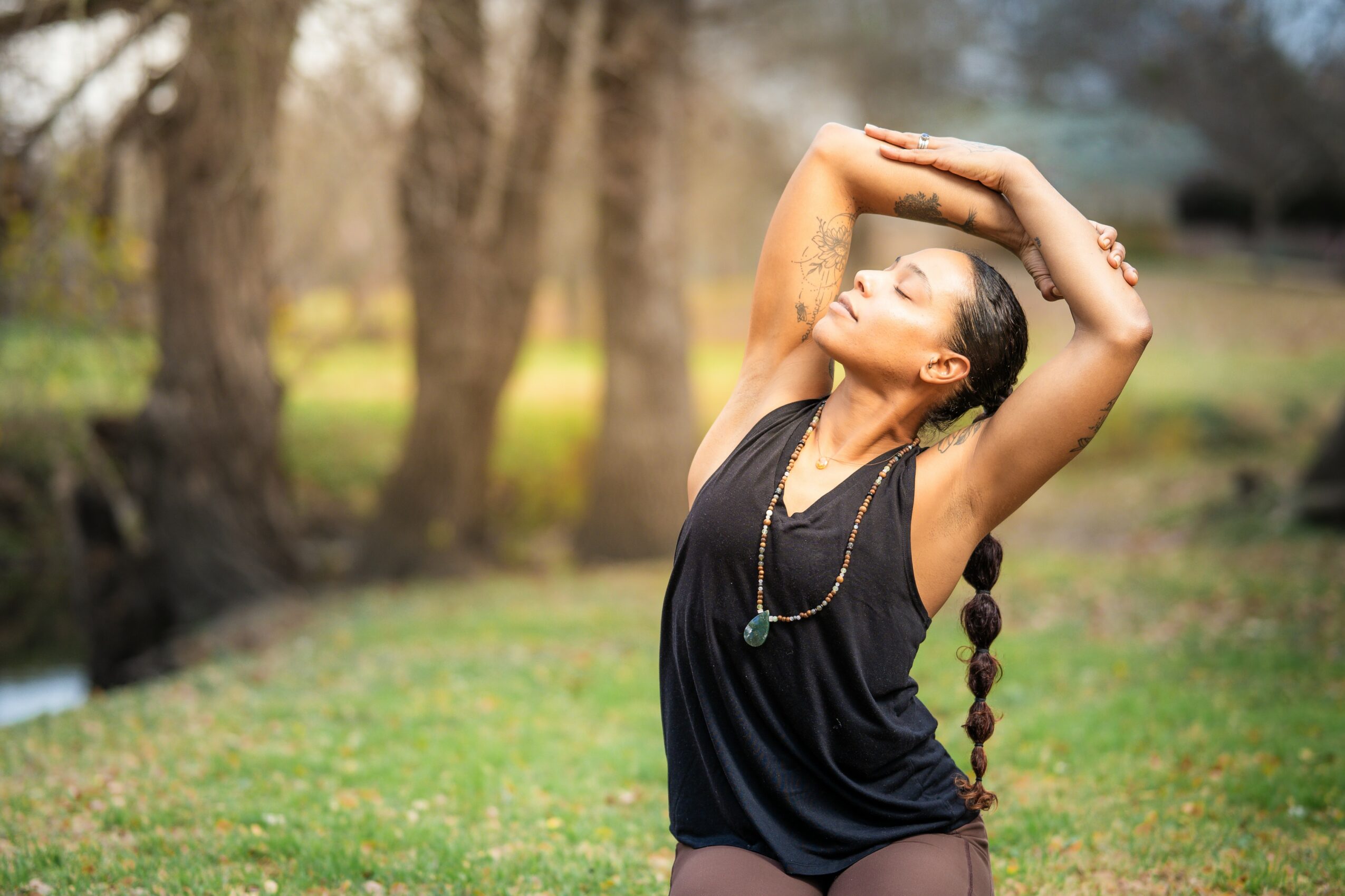 Woman with braided hair stretching outdoors in a park, trees in background, wearing a black sleeveless top, eyes closed.