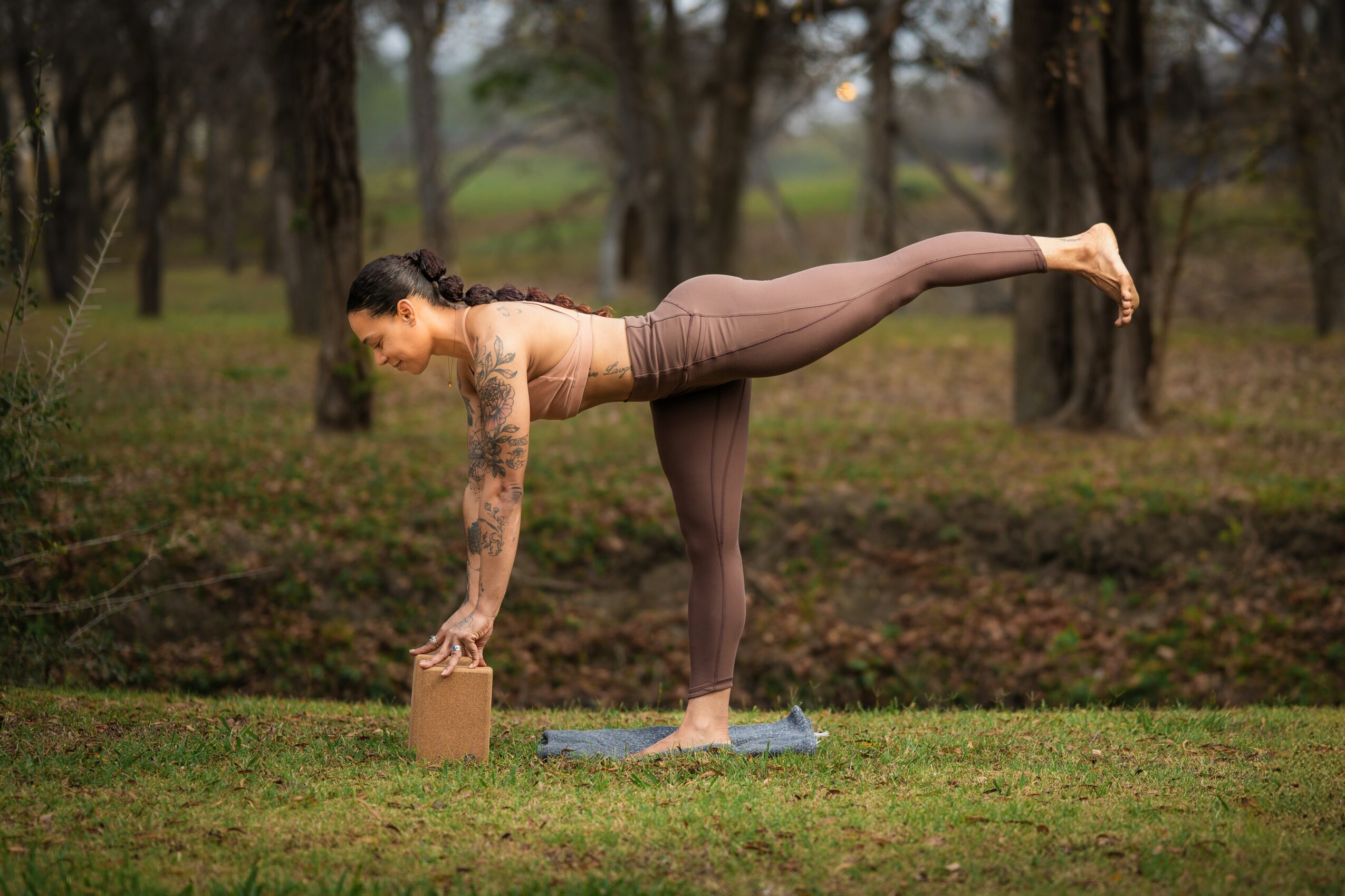 Woman practicing yoga outdoors, balancing on one leg with the other leg extended back, holding a brick for support, in a park.